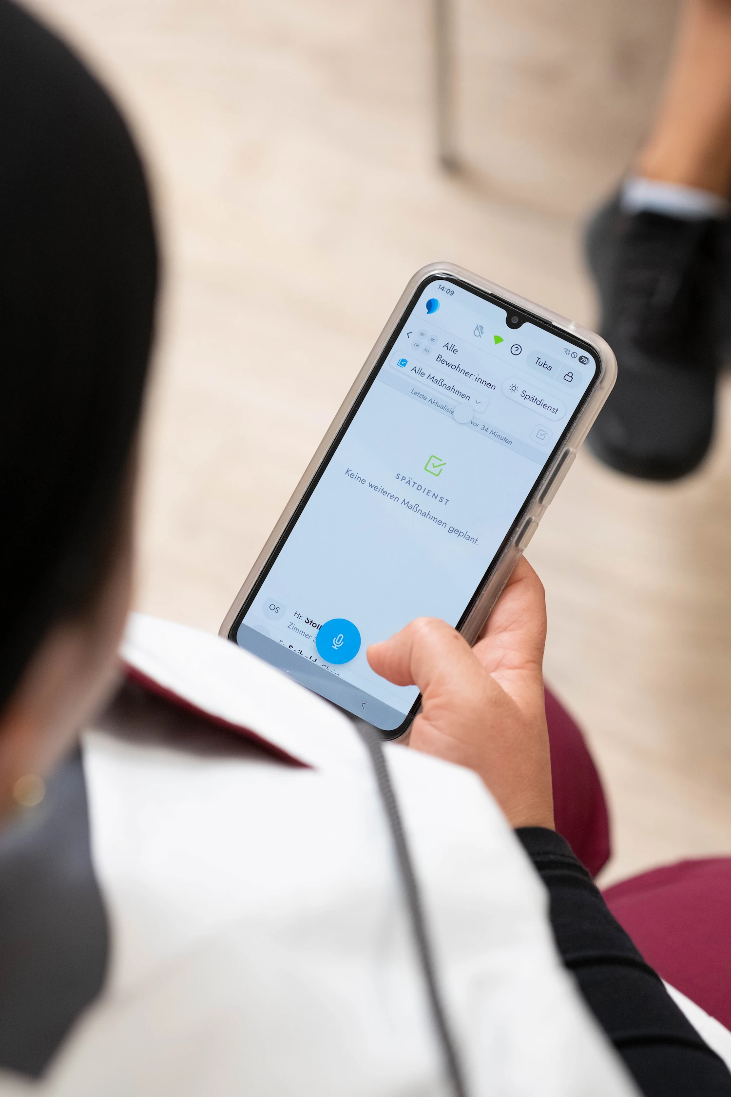Eine Pflegekraft hält ein Smartphone mit der voize Pflegedokumentations-App, auf der der Status „Spätdienst“ und ein blaues Mikrofon-Symbol für die sprachgestützte Eingabe zu sehen sind.