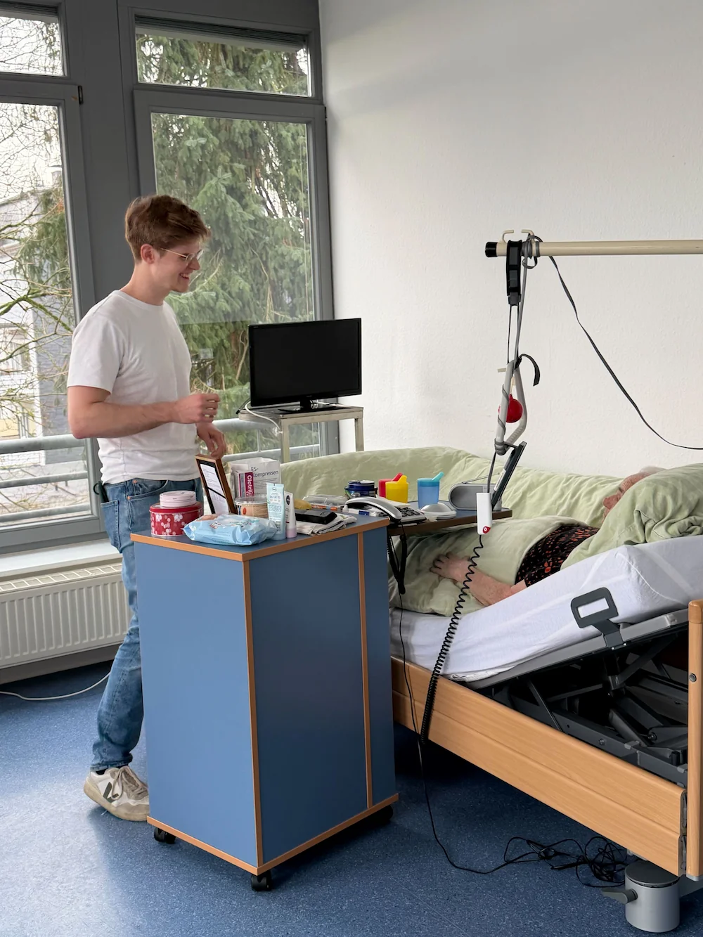 Junger Pfleger lächelt einen älteren Patienten an, der in einem hellen Pflegezimmer im Bett liegt.
