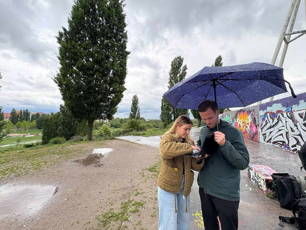 Zwei voize-Teammitglieder arbeiten gemeinsam an einem Laptop im Freien unter einem blauen Regenschirm in einem Park.