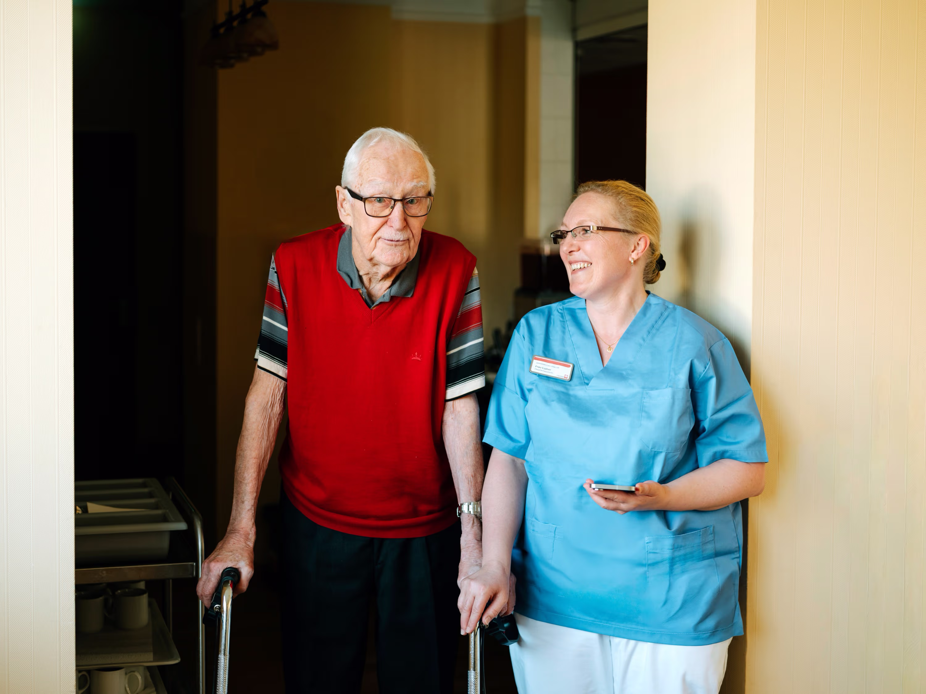 Älterer Mann mit Gehhilfe und Krankenschwester in blauer Uniform lachen zusammen.