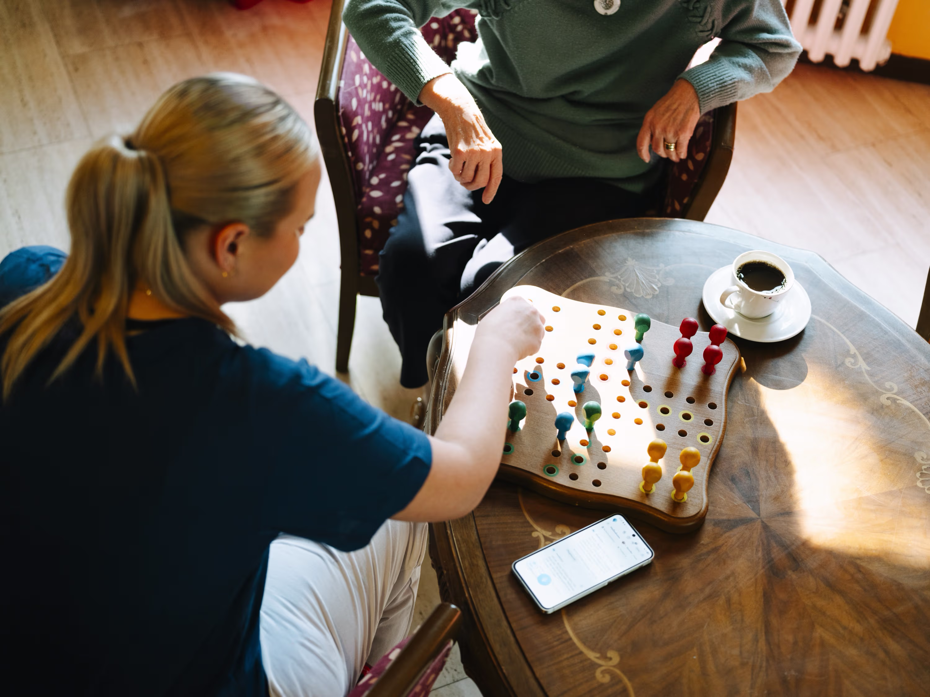 Junge Frau und ältere Person spielen ein Brettspiel mit bunten Stiften an einem Holztisch.