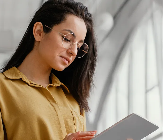 Woman with glasses in a yellow shirt reading or using a tablet near a bright window.