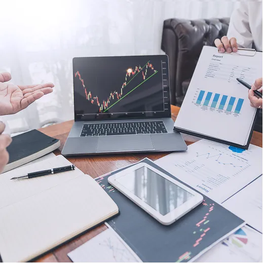 Person showing financial report with charts beside laptop displaying stock market graph on wooden table.
