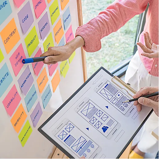 Two people planning a website layout with sticky notes on board and a wireframe on clipboard.