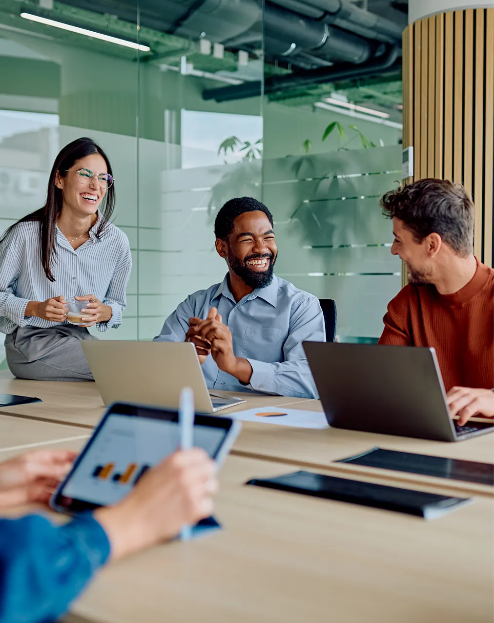 Three coworkers smiling and talking during a meeting in a glass-walled office with laptops.