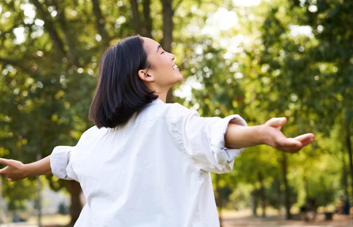 Woman with outstretched arms looking up and smiling in a green, sunny park.