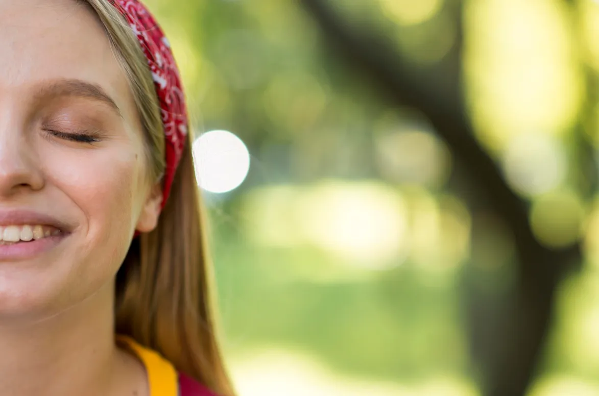 Close-up of a smiling woman with closed eyes wearing a red headband outdoors with blurred green background.
