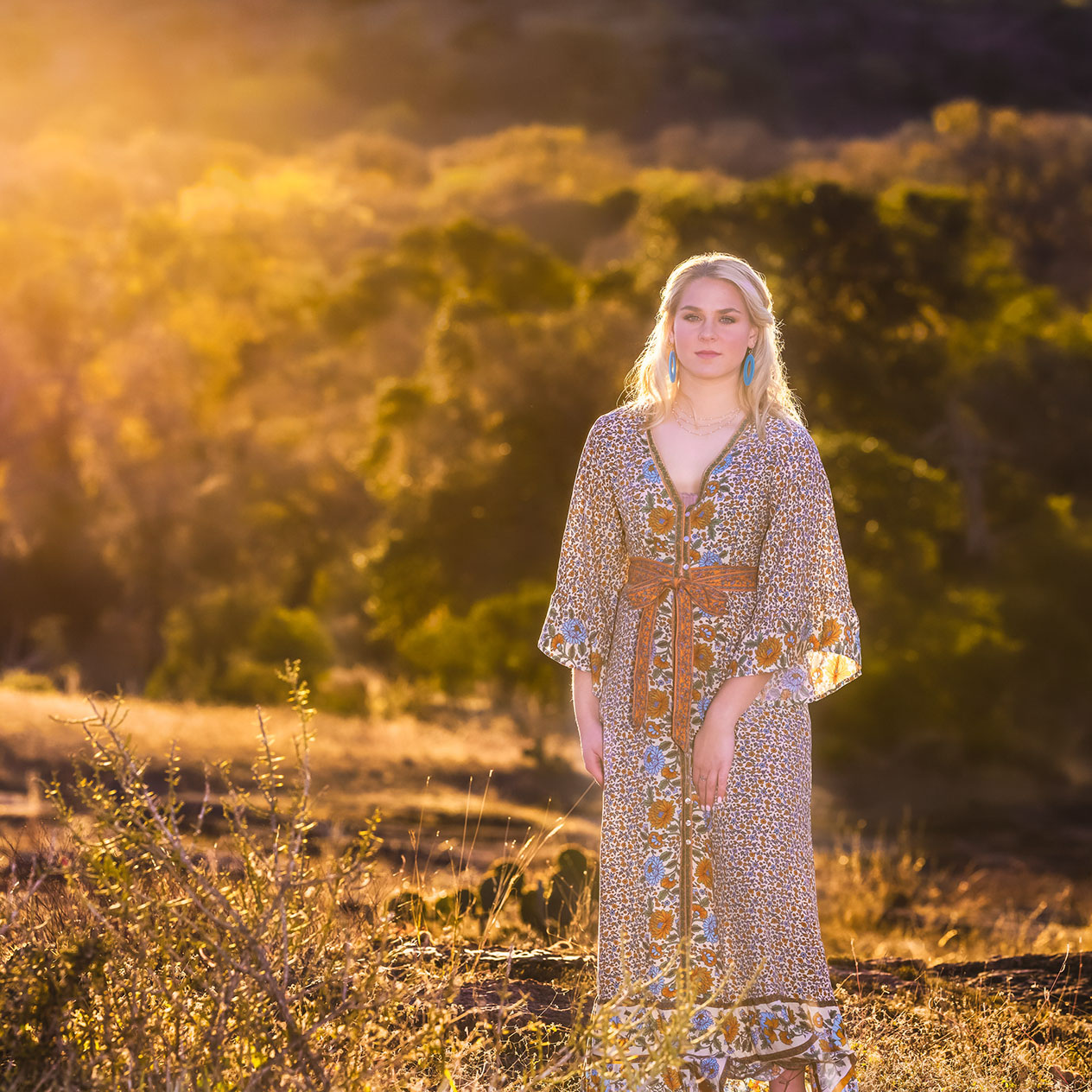 Blonde woman wearing a long floral dress and blue earrings standing in a sunlit field with trees in the background.