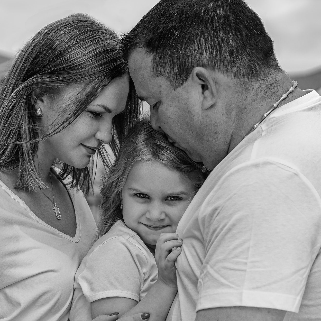 Black and white photo of a mother and father lovingly embracing their smiling young daughter between them.