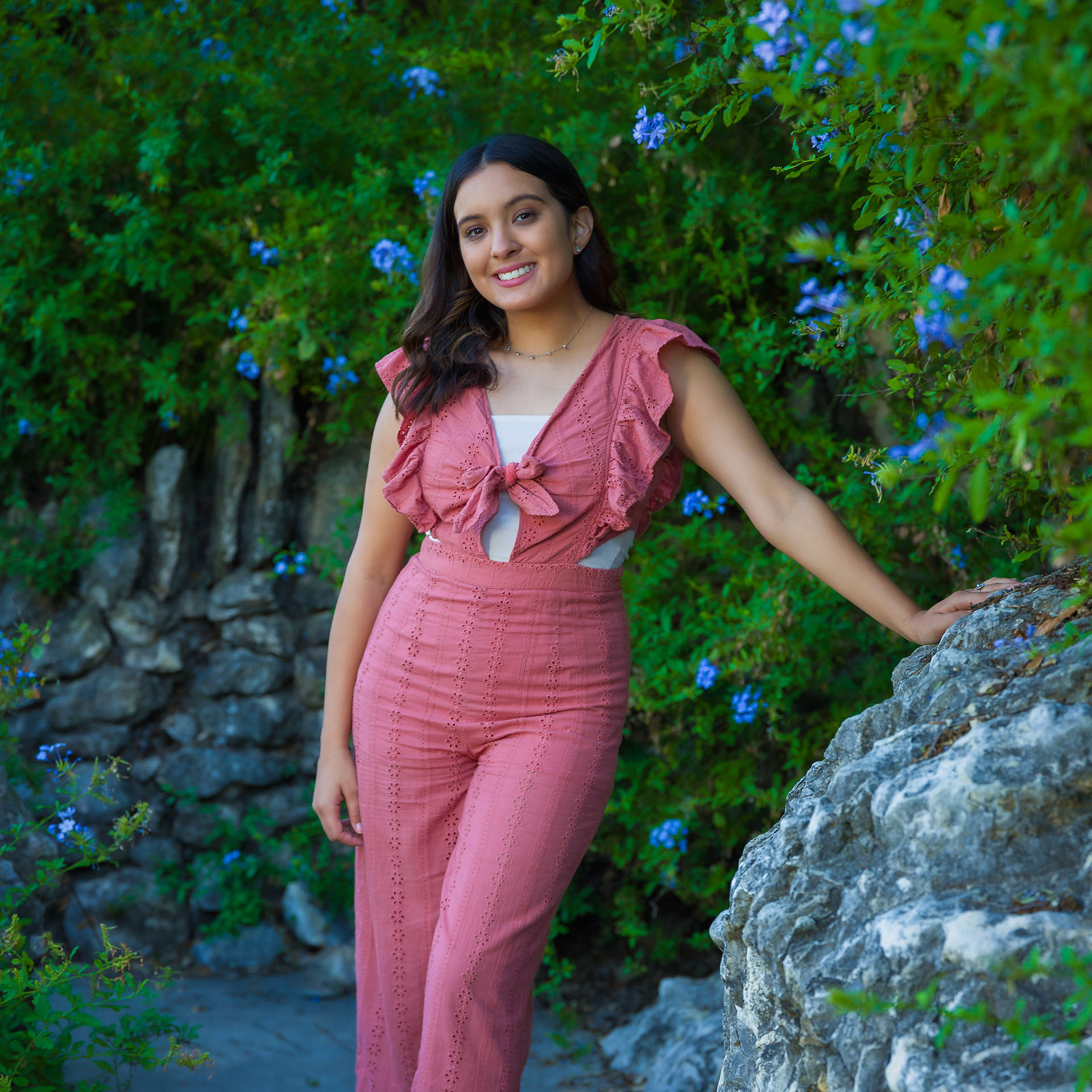 Young woman in a pink ruffled jumpsuit smiling and leaning on a rock wall surrounded by green foliage with blue flowers.