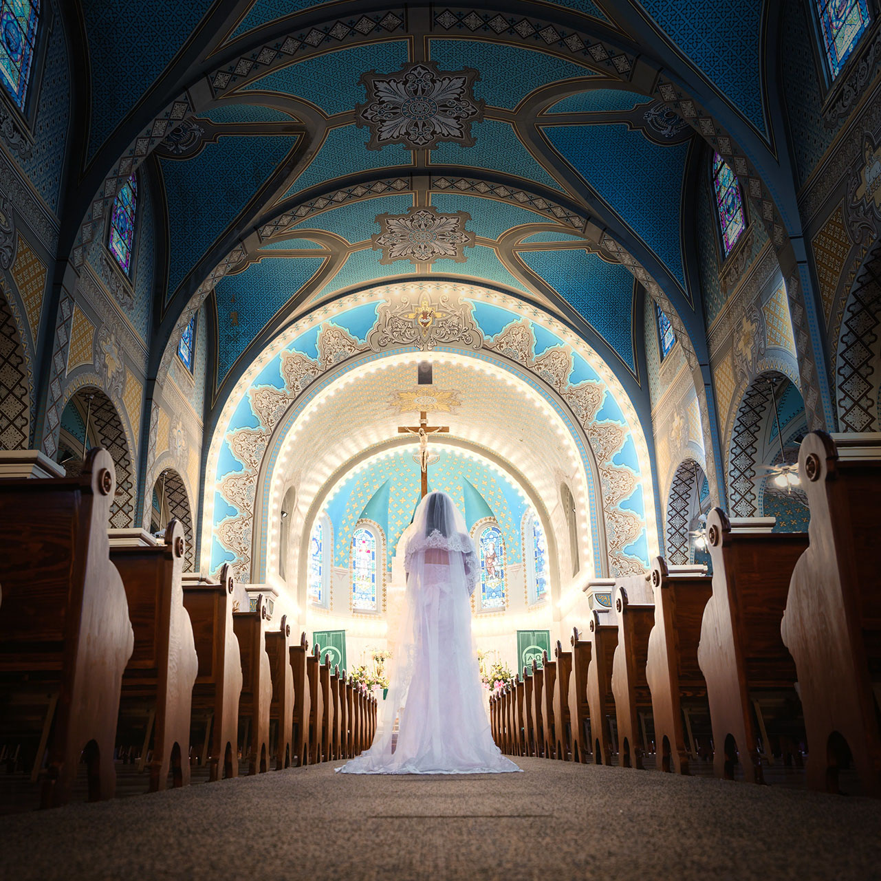 Woman in a white wedding dress standing in the aisle of a church with ornate blue ceiling and stained glass windows.