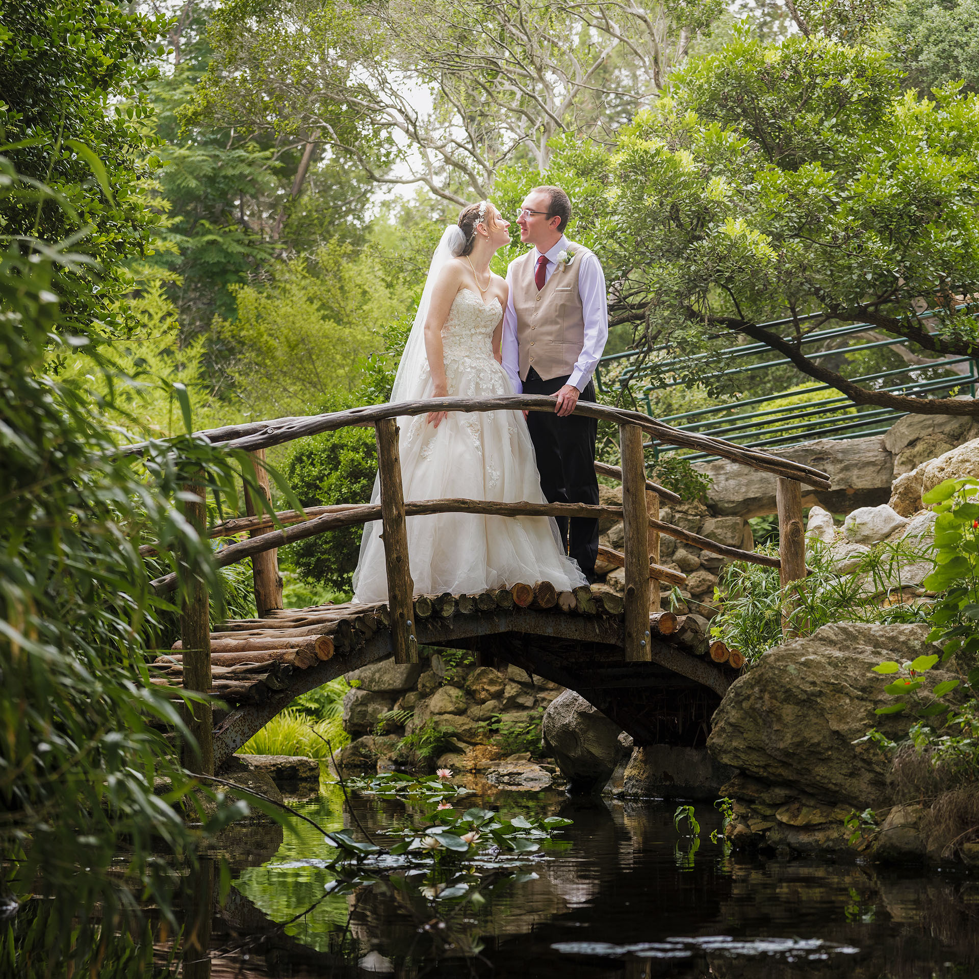 Bride and groom standing on a rustic wooden bridge over a pond in a lush green garden.