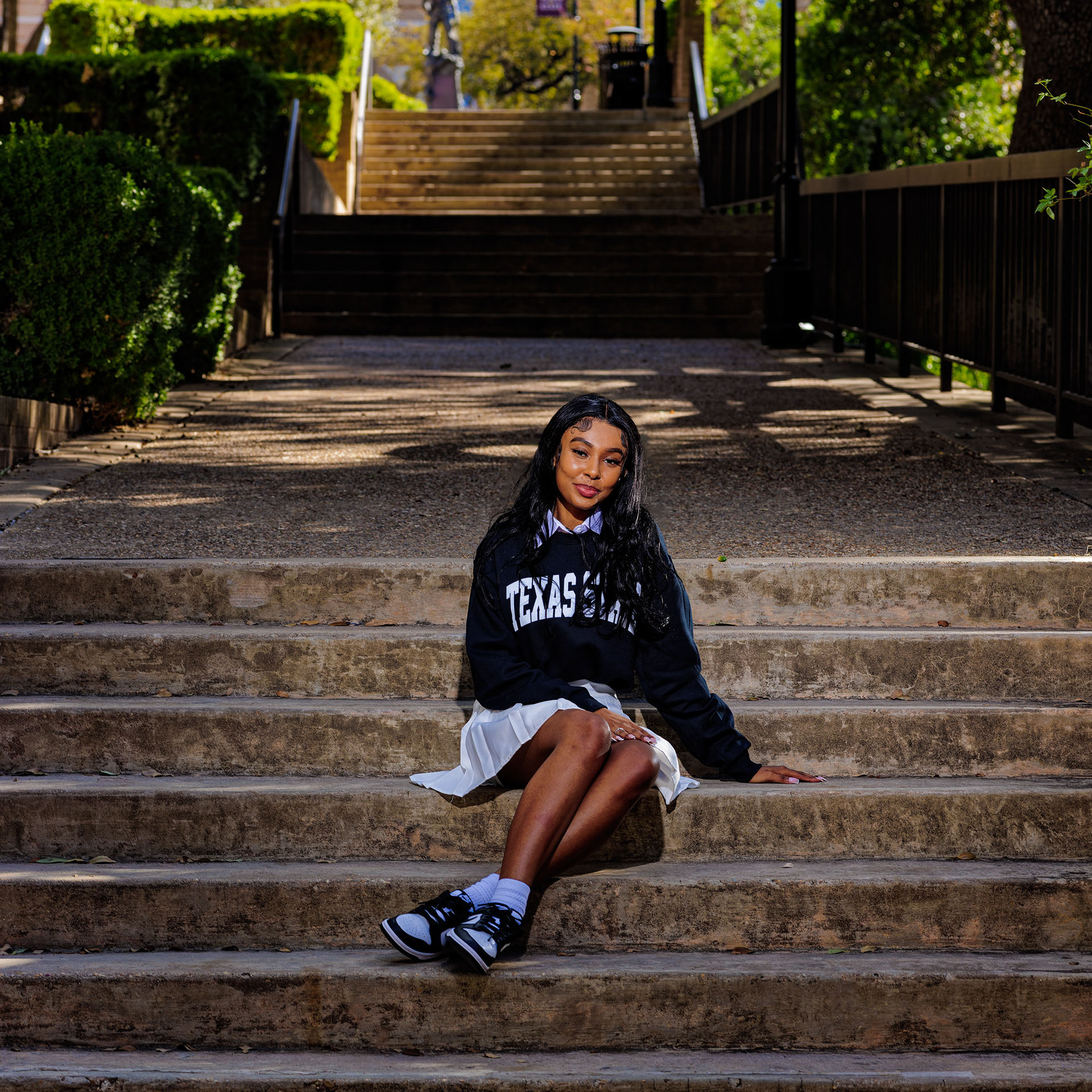 Smiling young woman sitting on stone steps wearing a Texas State sweatshirt and white skirt.