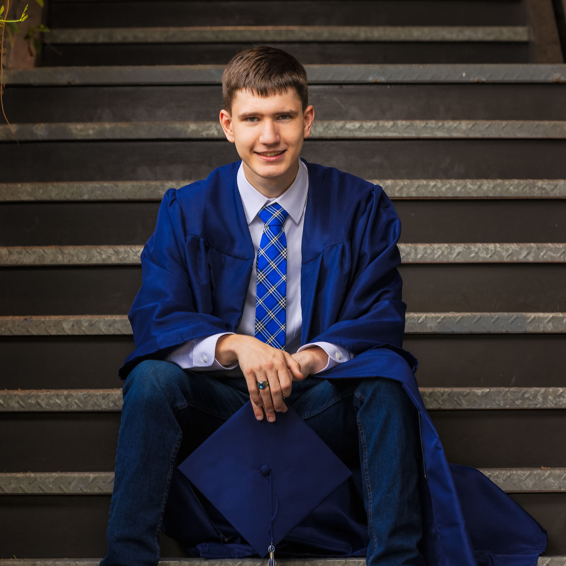Young man in a blue graduation gown and tie sitting on stairs holding a graduation cap.