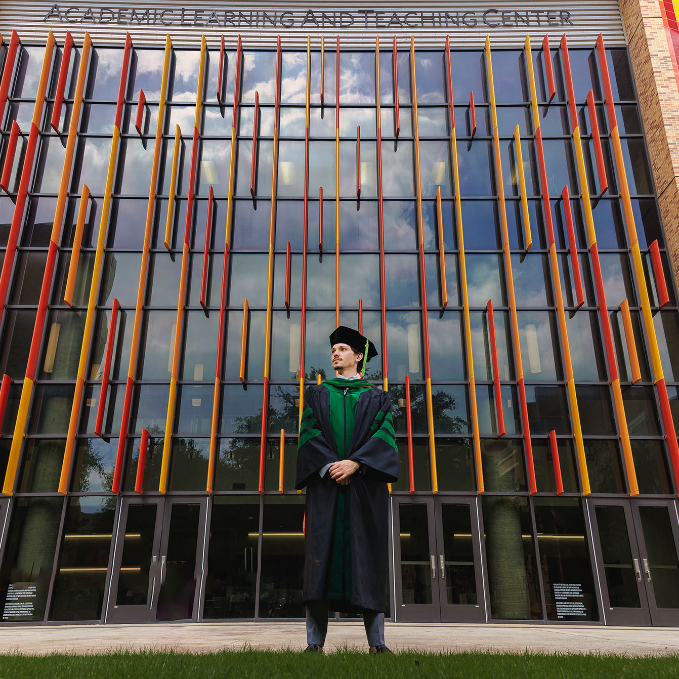 Graduate in black cap and gown with green robes standing confidently in front of a modern glass building with vertical red and yellow architectural accents under the sign Academic Learning and Teaching Center.
