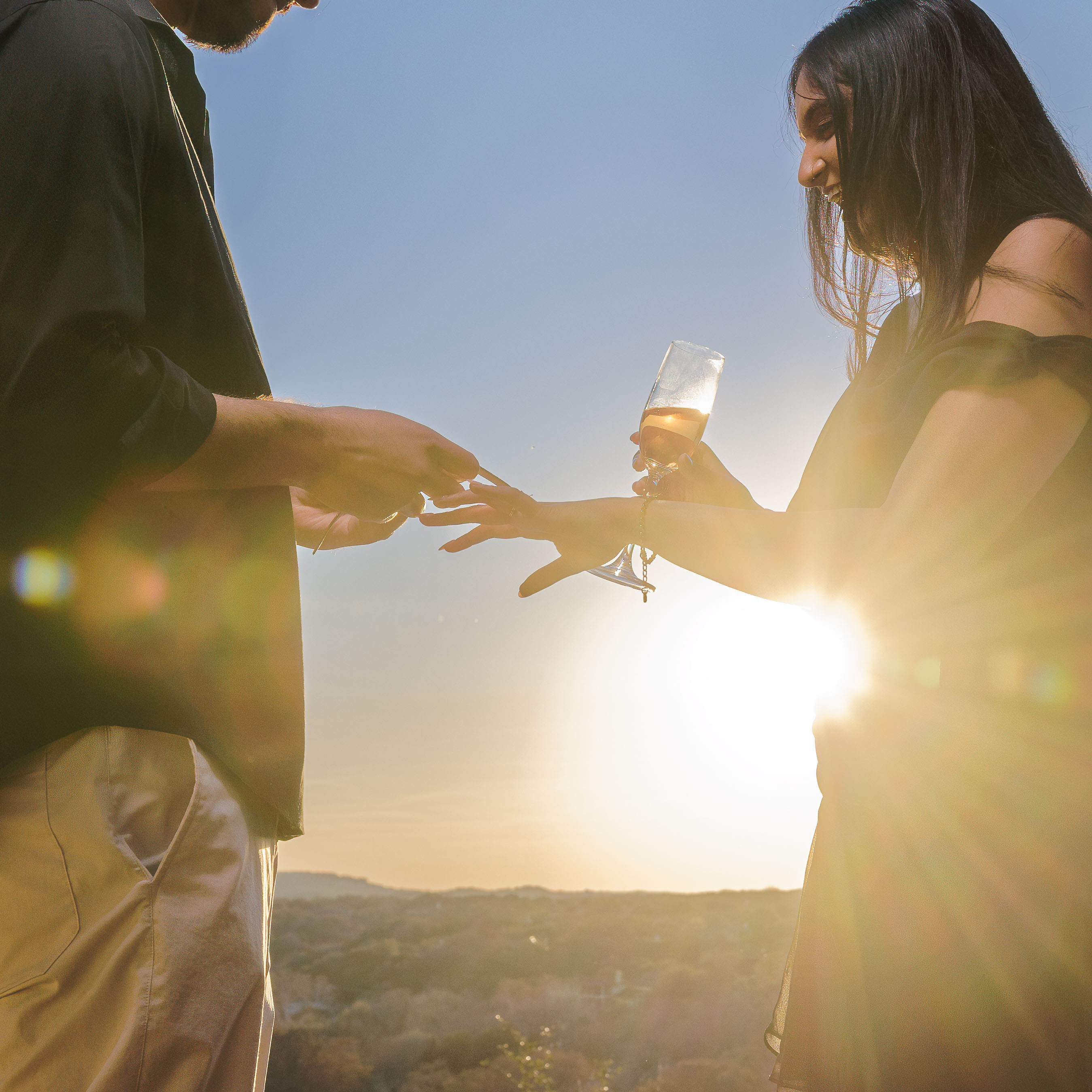 Man placing a ring on woman's finger as she holds a glass of champagne at sunset outdoors.