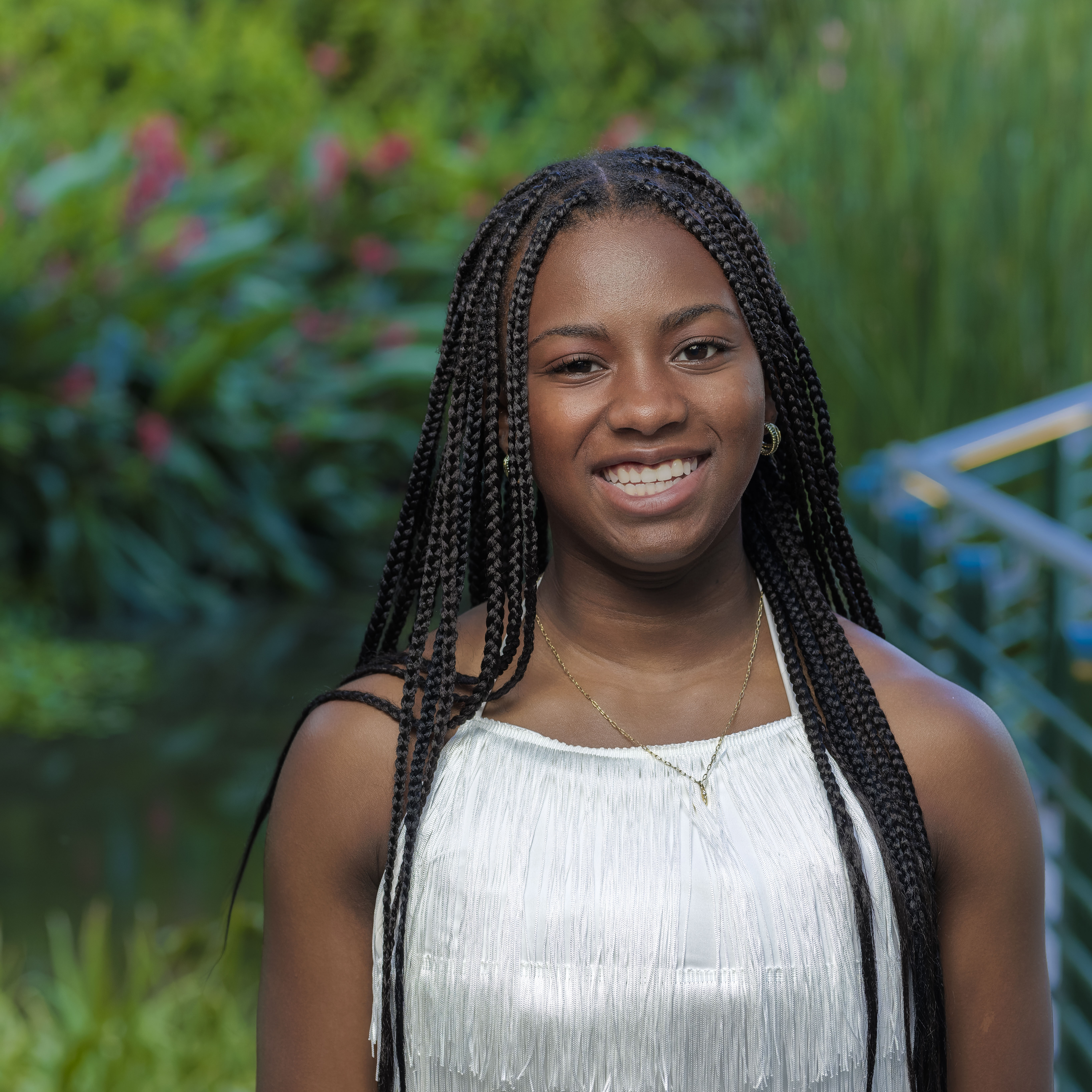 Smiling young woman with long braided hair wearing a white sleeveless top, standing outdoors with greenery in the background.