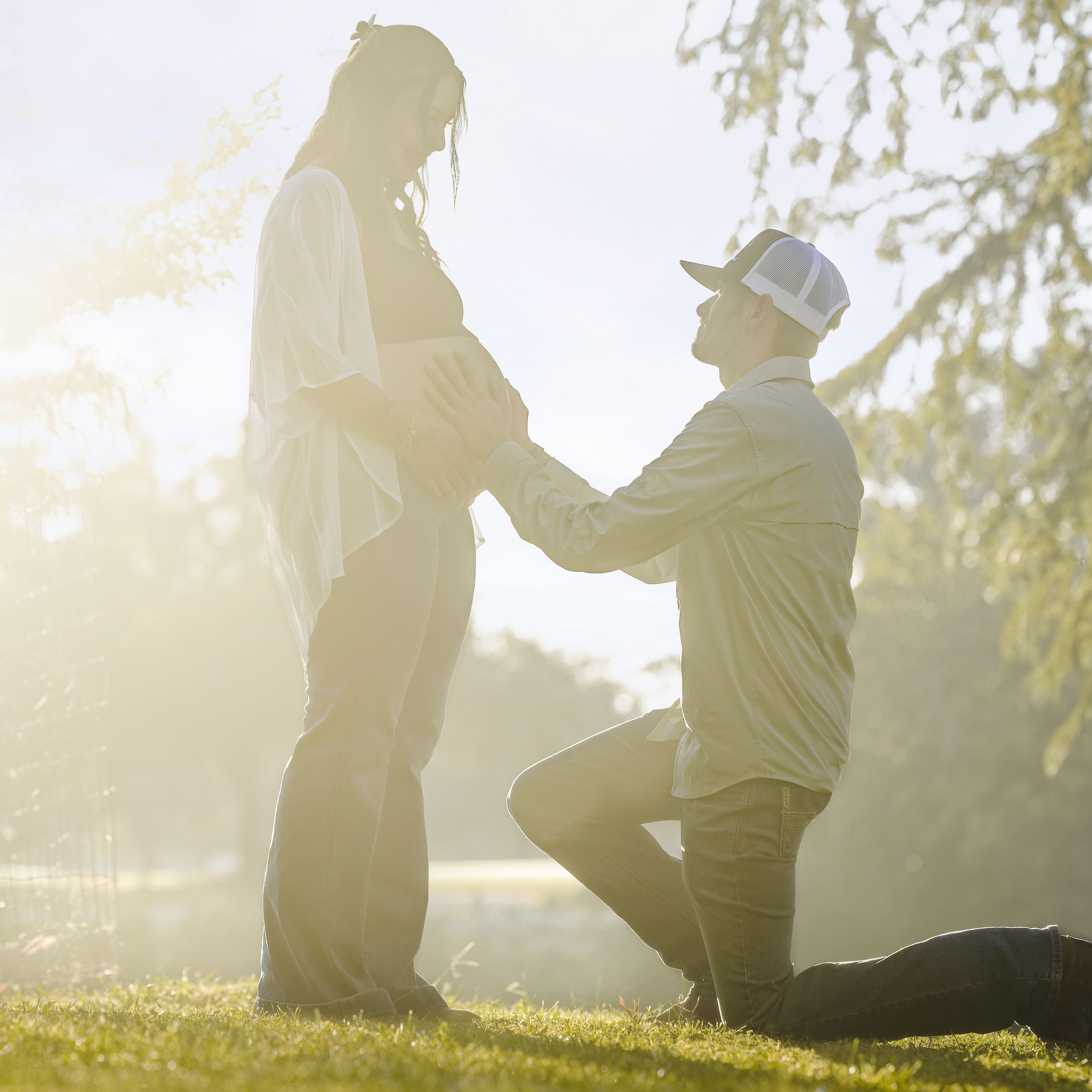 Man kneeling on one knee holding the belly of a pregnant woman standing in a sunlit outdoor setting.