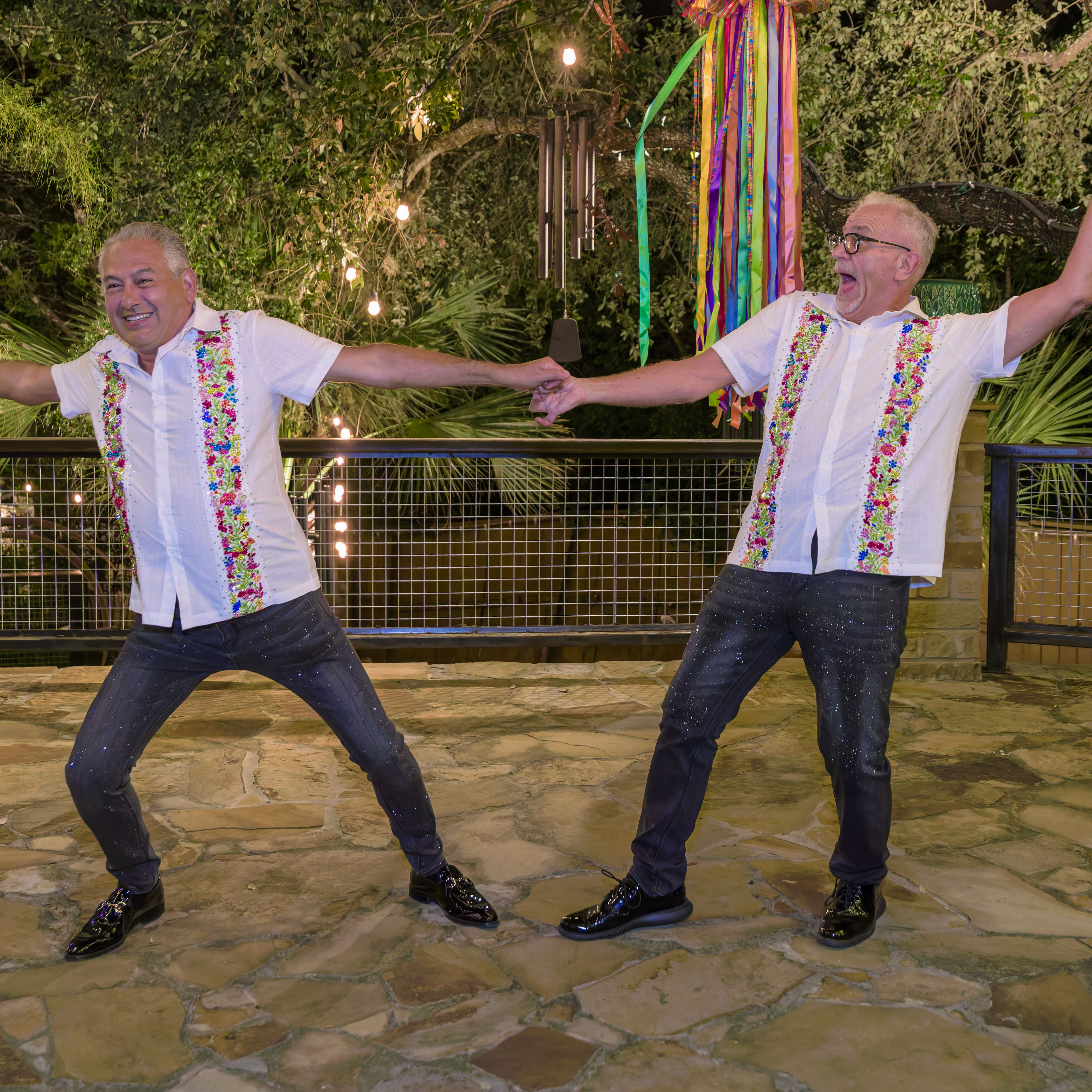 Two men holding hands and dancing outdoors at night, wearing white shirts with colorful floral embroidery and dark pants.