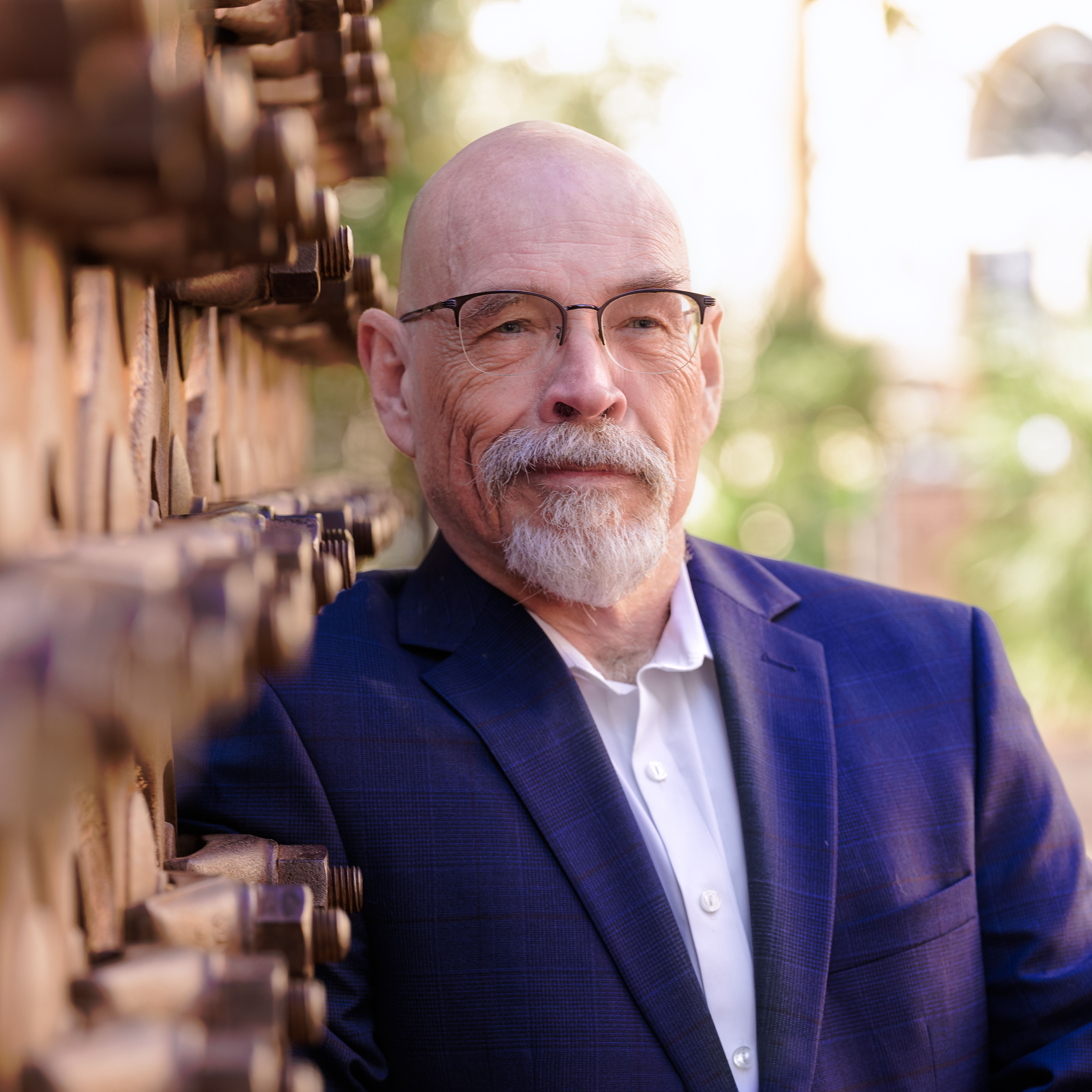 Portrait of a serious bald man with glasses and a white beard wearing a blue suit jacket and white shirt, leaning against a rusty metal wall outdoors.