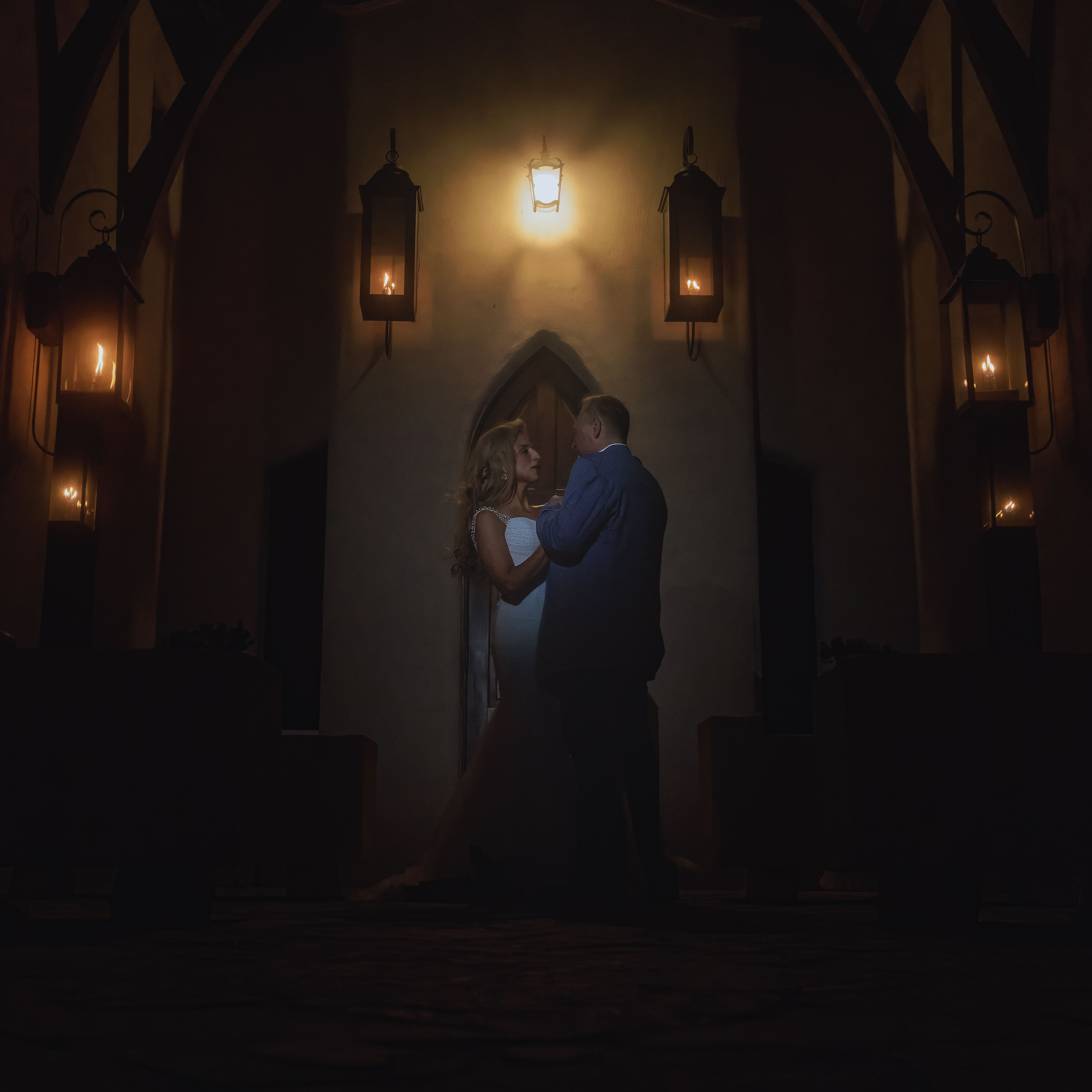 Bride in white wedding gown and groom in suit holding hands and facing each other in a dimly lit room with wall lanterns.