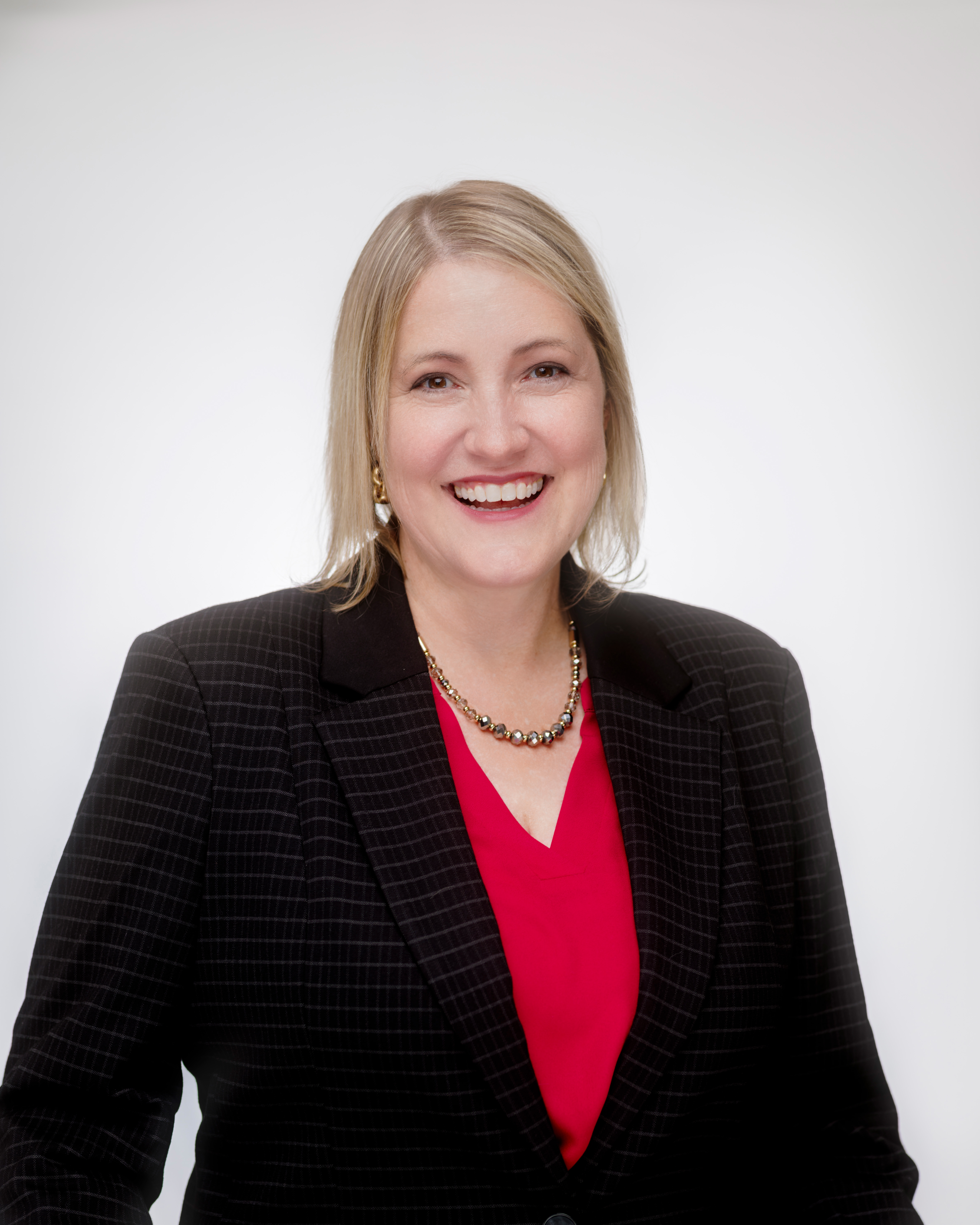 Smiling woman with blonde hair wearing a black striped blazer and red blouse with necklace.