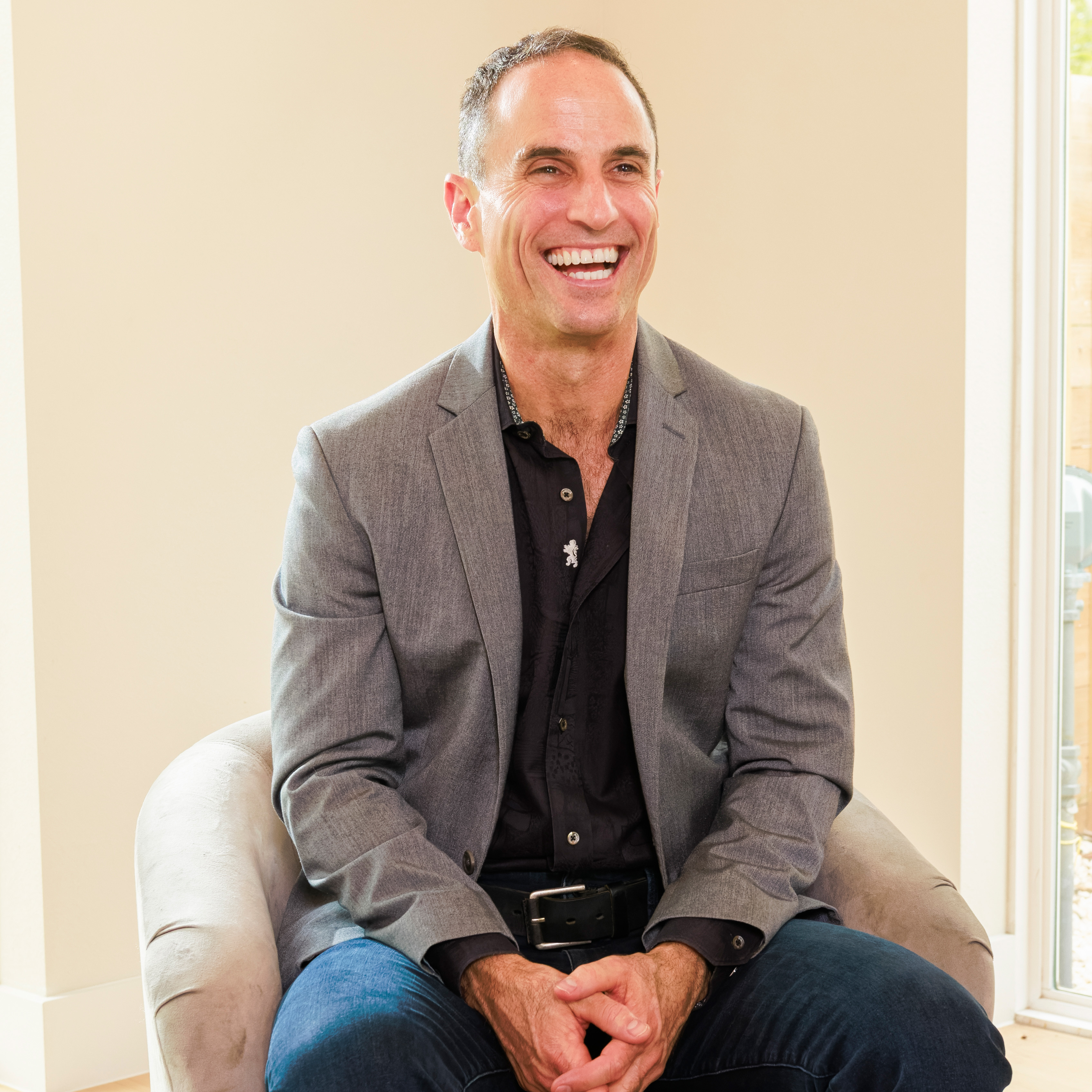 Smiling man in a gray blazer and black shirt sitting on a beige chair near a window.