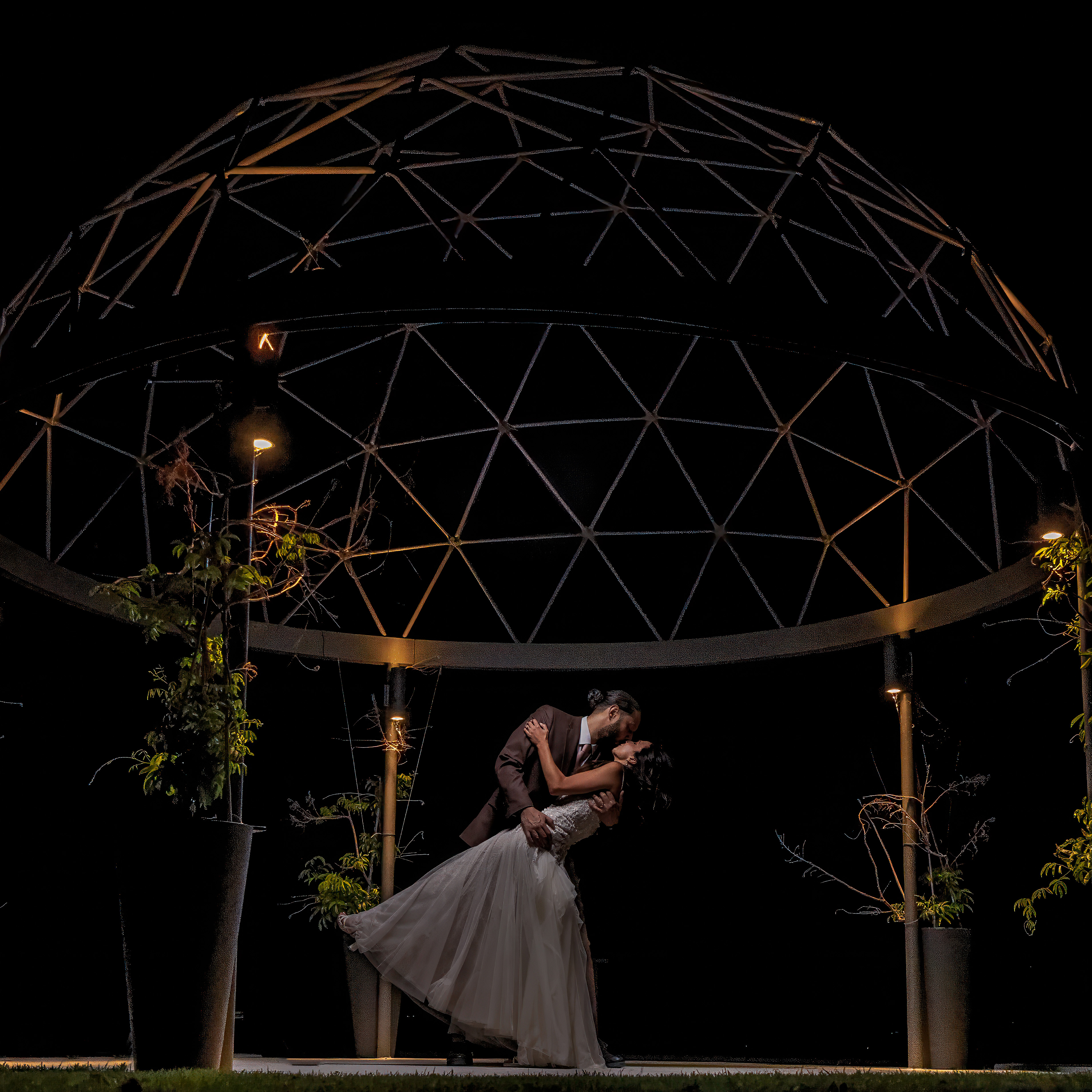 Bride and groom kissing with the groom dipping the bride under a lit metal dome at night.