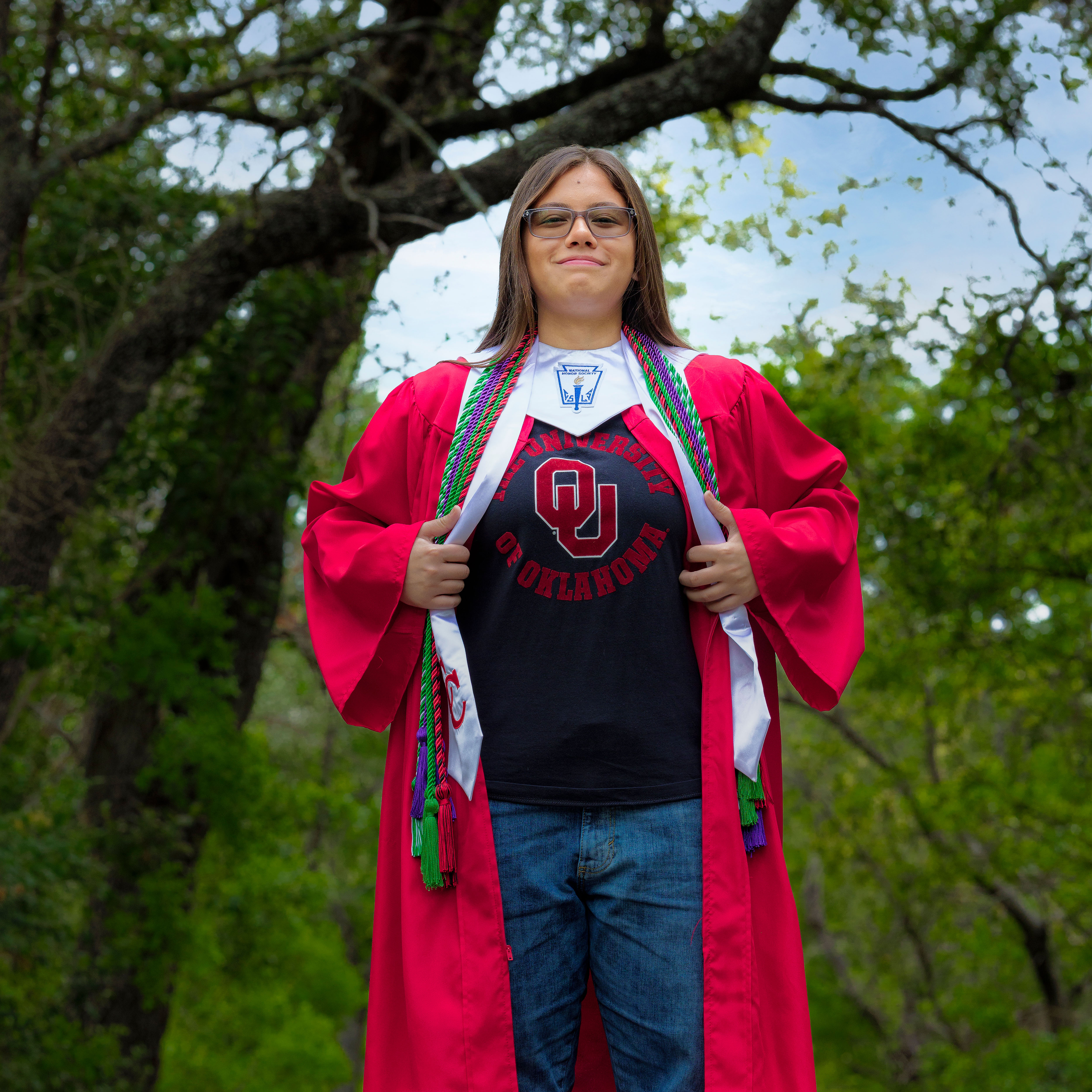 Graduate in red gown and cords showing University of Oklahoma shirt outside with trees behind.