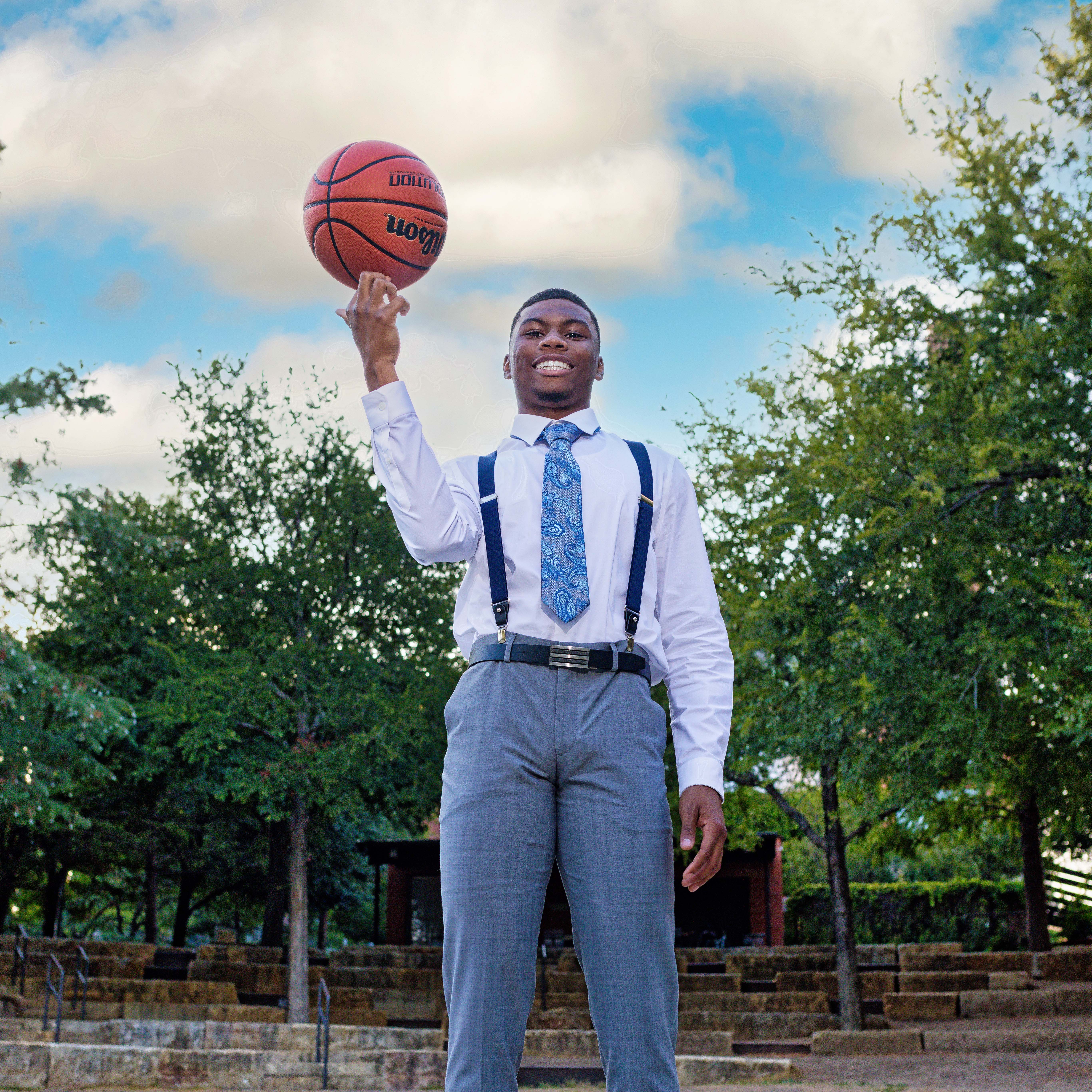 Young man in dress shirt, tie, suspenders spinning a basketball on his finger outdoors.