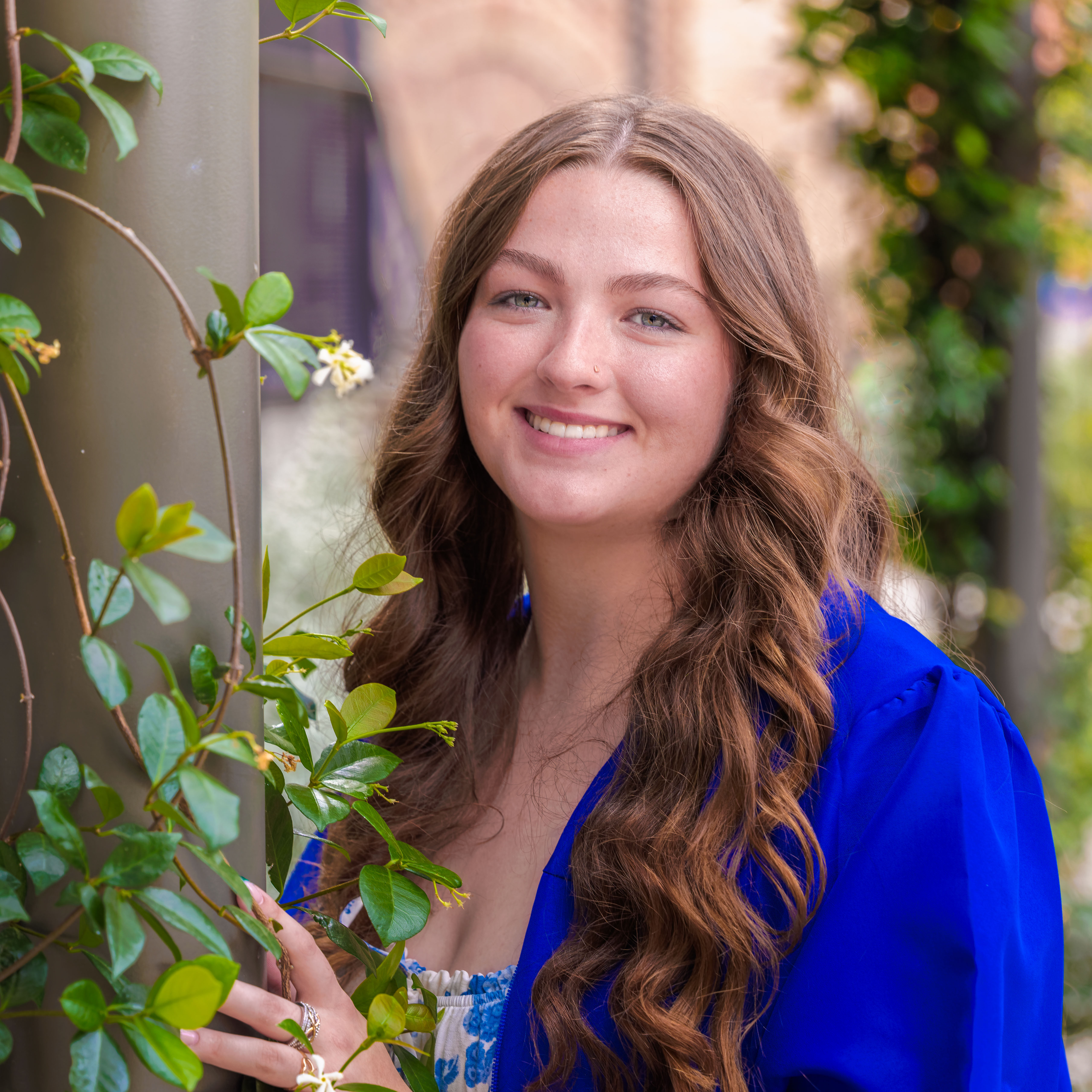 Young woman with long wavy brown hair smiling, wearing a blue graduation gown outdoors by greenery.