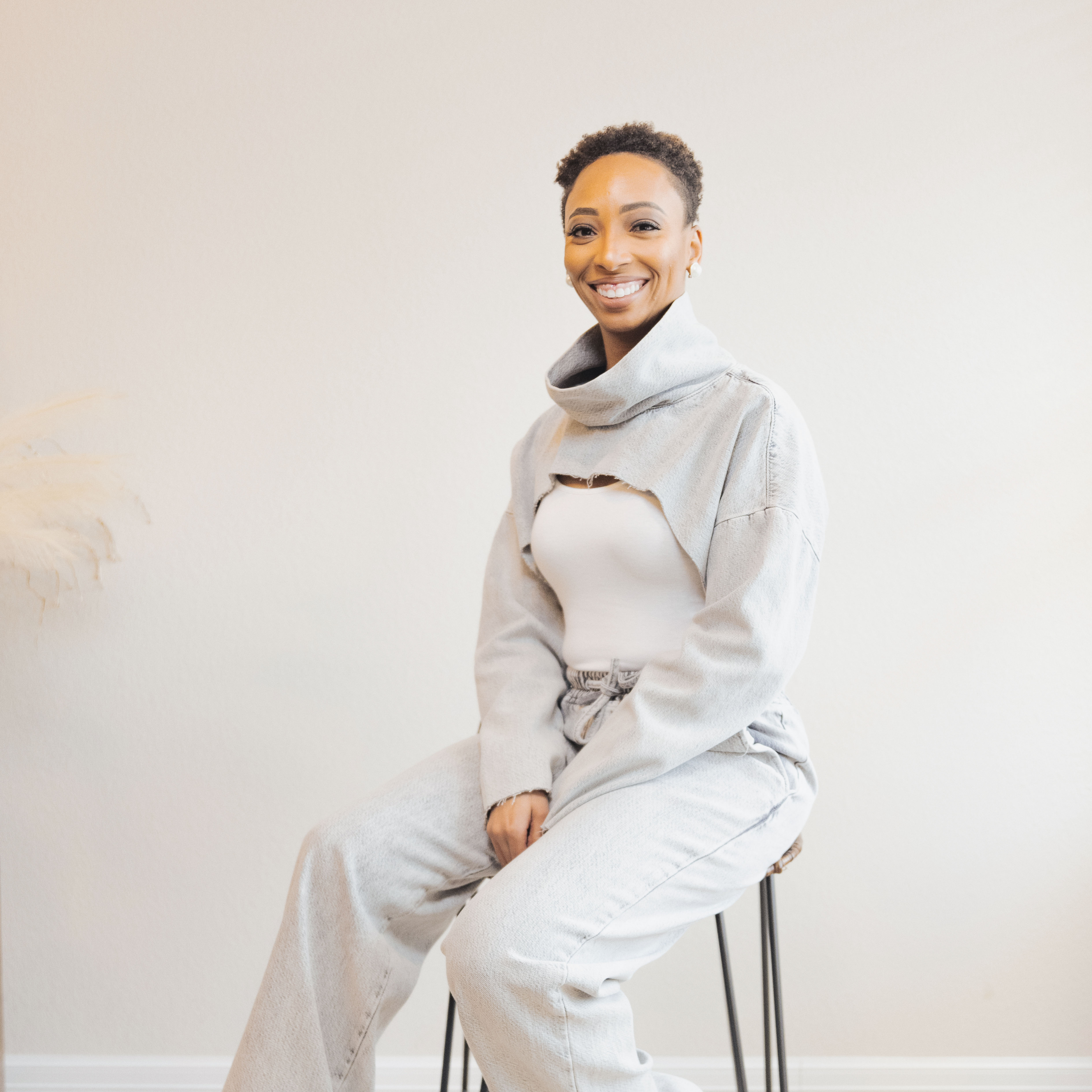 Smiling woman in light casual outfit sitting on tall stool against plain wall.