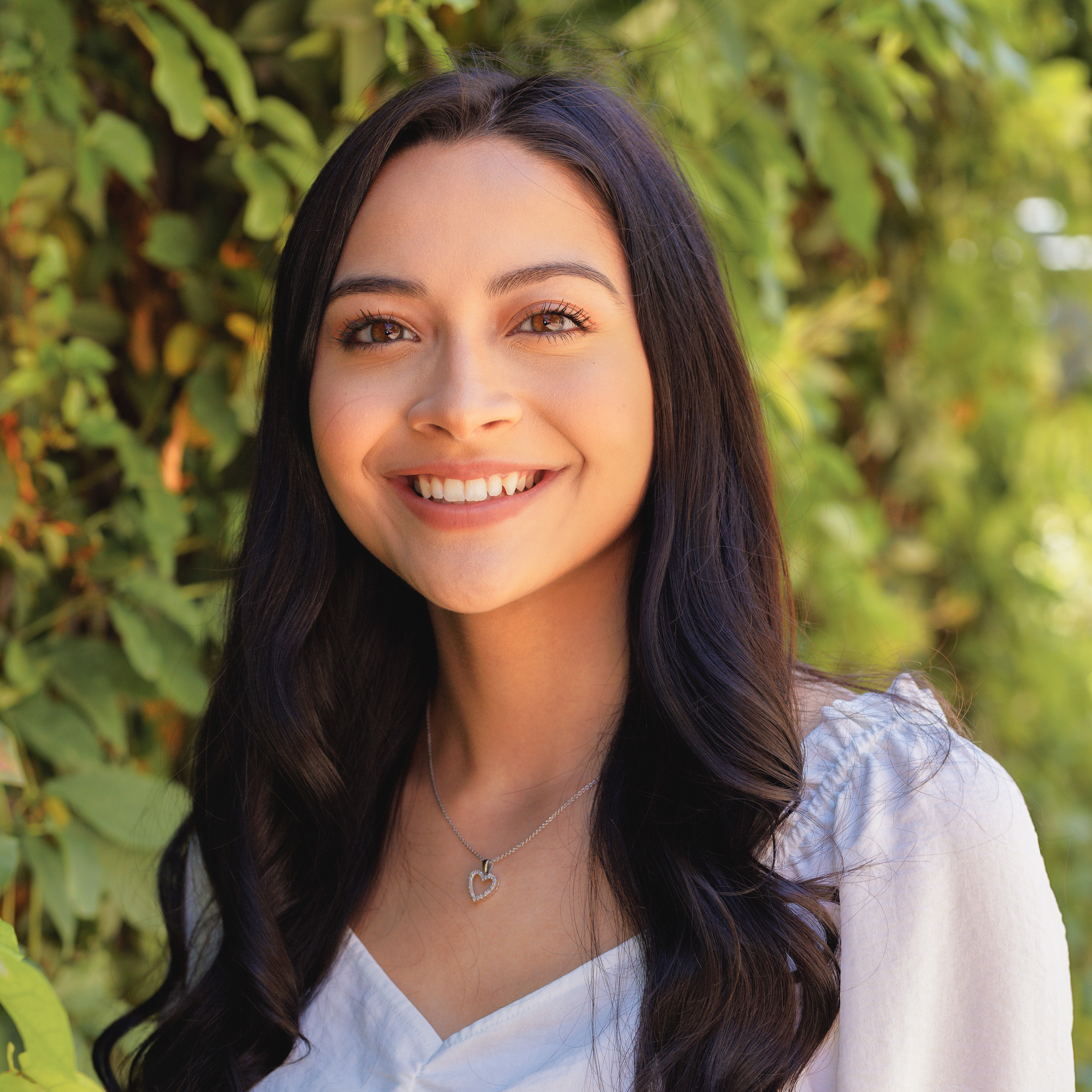 Smiling young woman with long dark hair and a white top stands before leafy green plants.