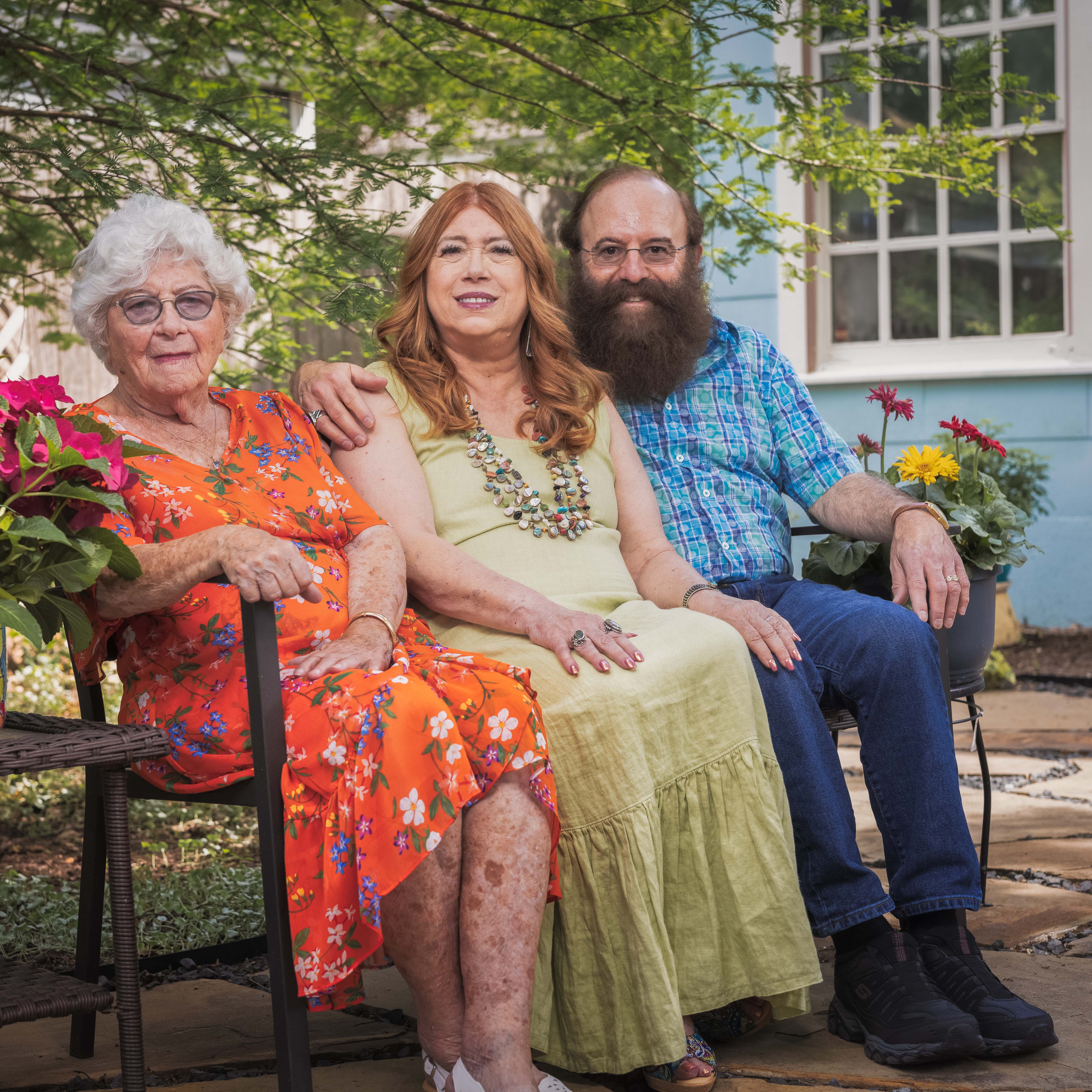 Three adults sitting on a bench outdoors with flowers and a house with windows behind them.