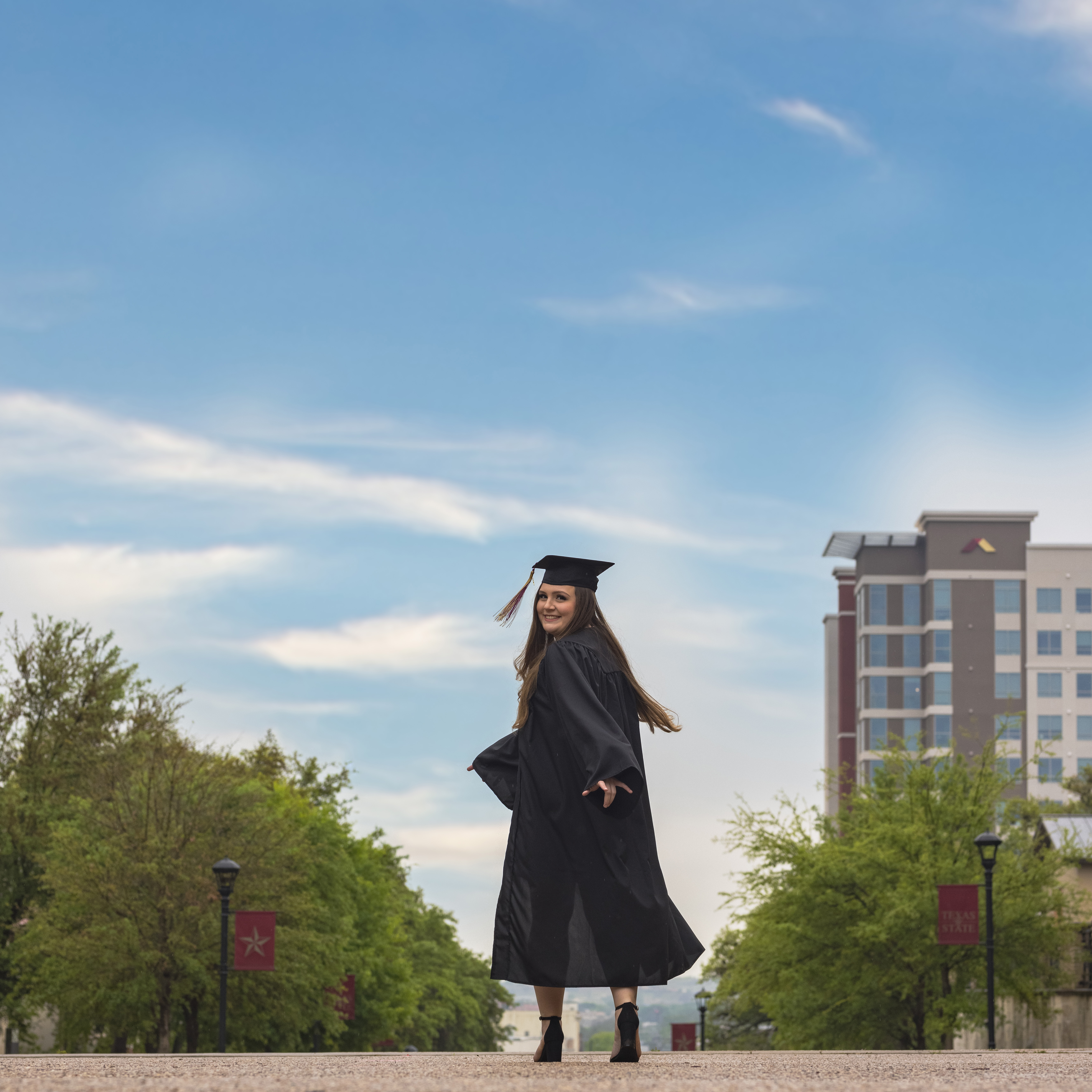 Smiling female graduate in cap and gown walking outdoors on a tree-lined street with buildings and blue sky.