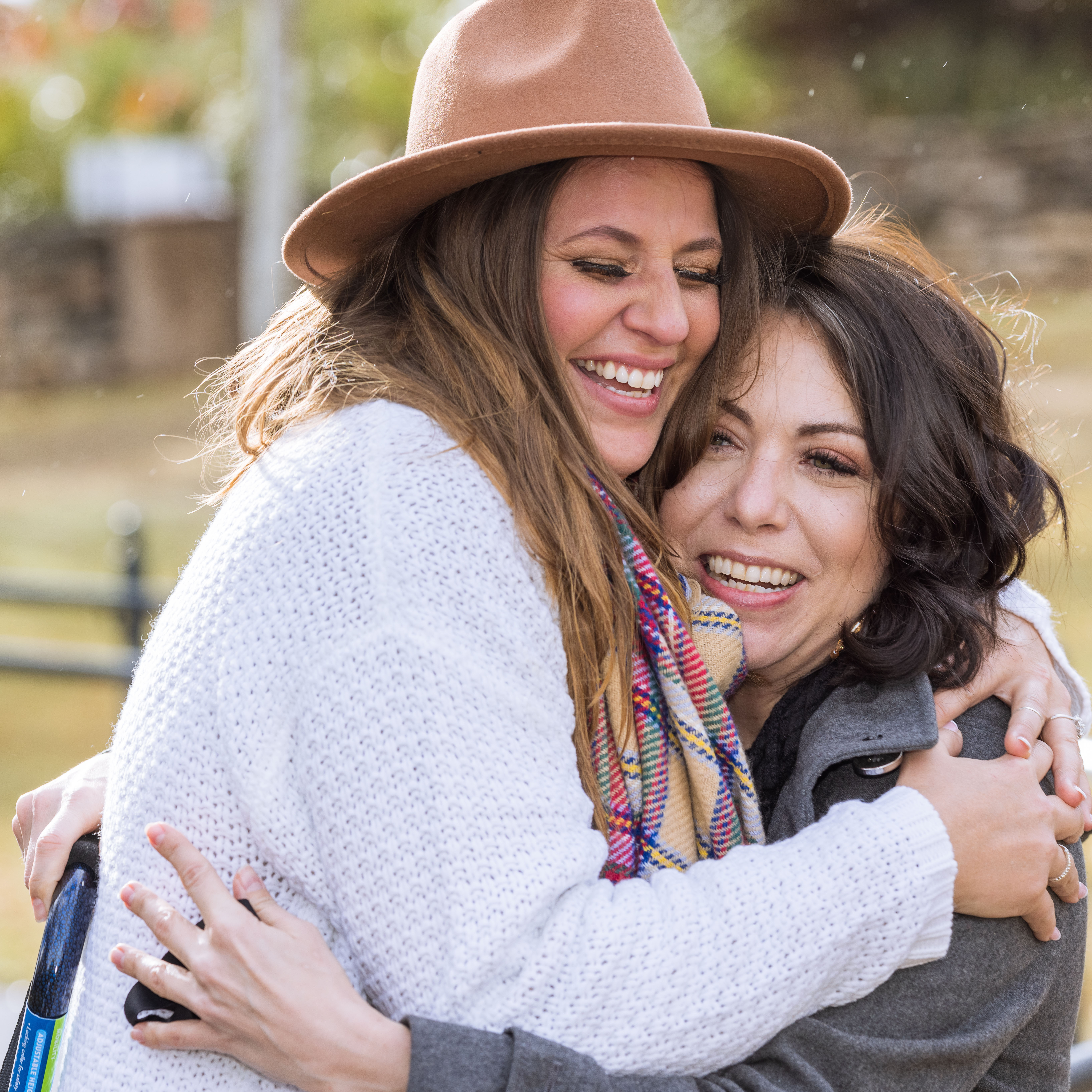 Two women warmly hugging outdoors, one wearing a brown hat and white sweater, both smiling joyfully.