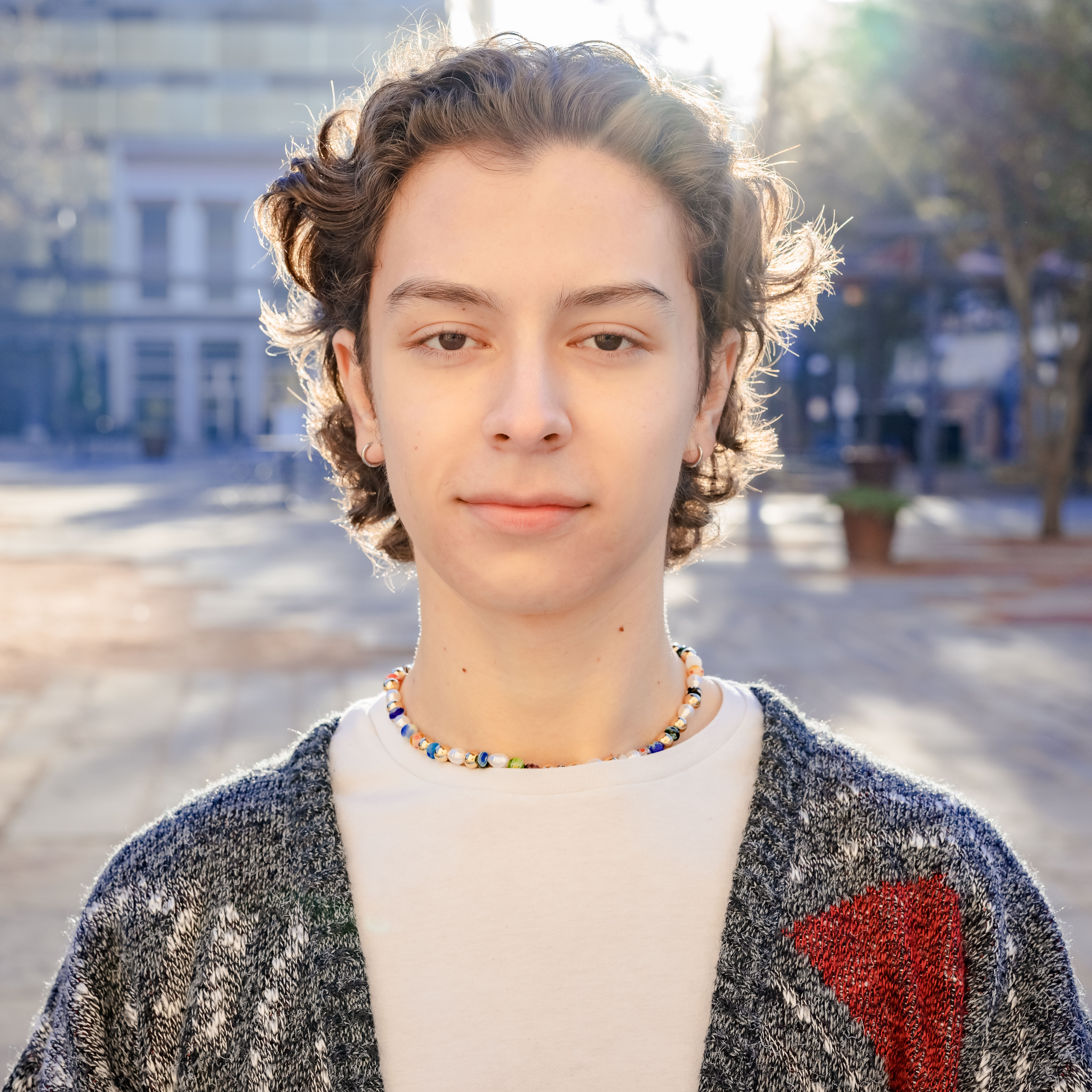 Young person with curly hair wearing a beaded necklace and knit cardigan outdoors in sunlight.
