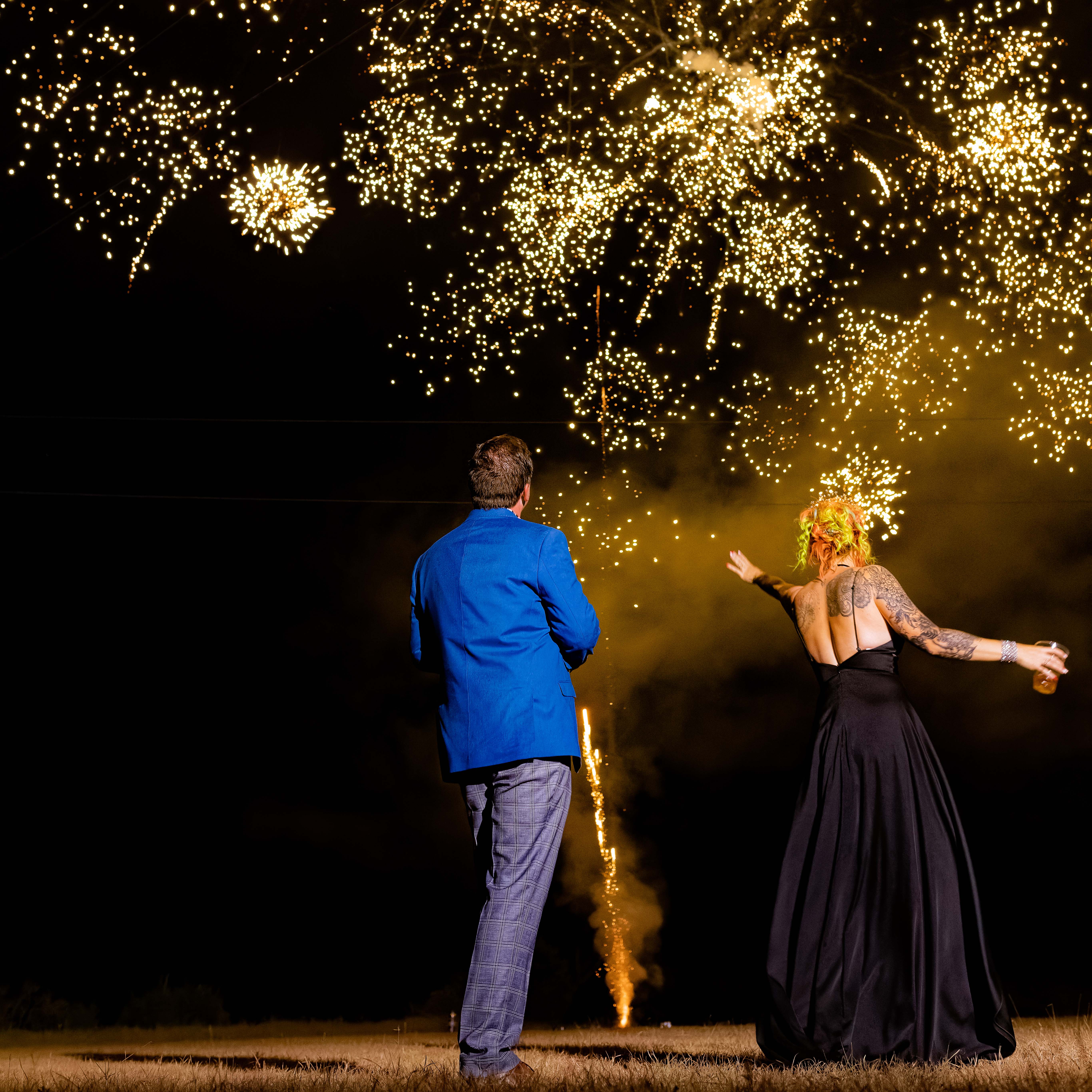 Man in blue suit and woman in black dress watch fireworks at night outdoors.