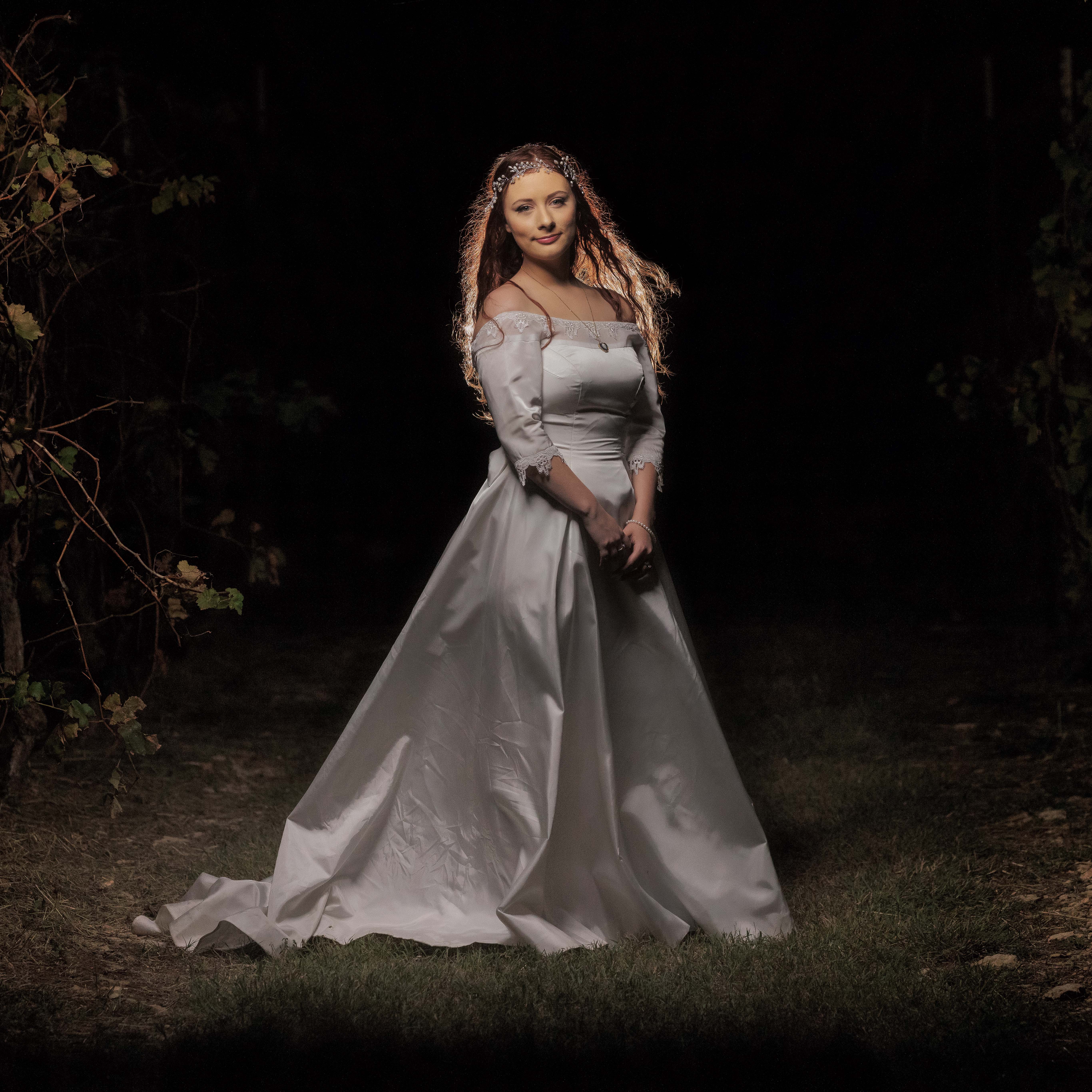 Woman in a long white dress with lace sleeves and headpiece standing outside at night.