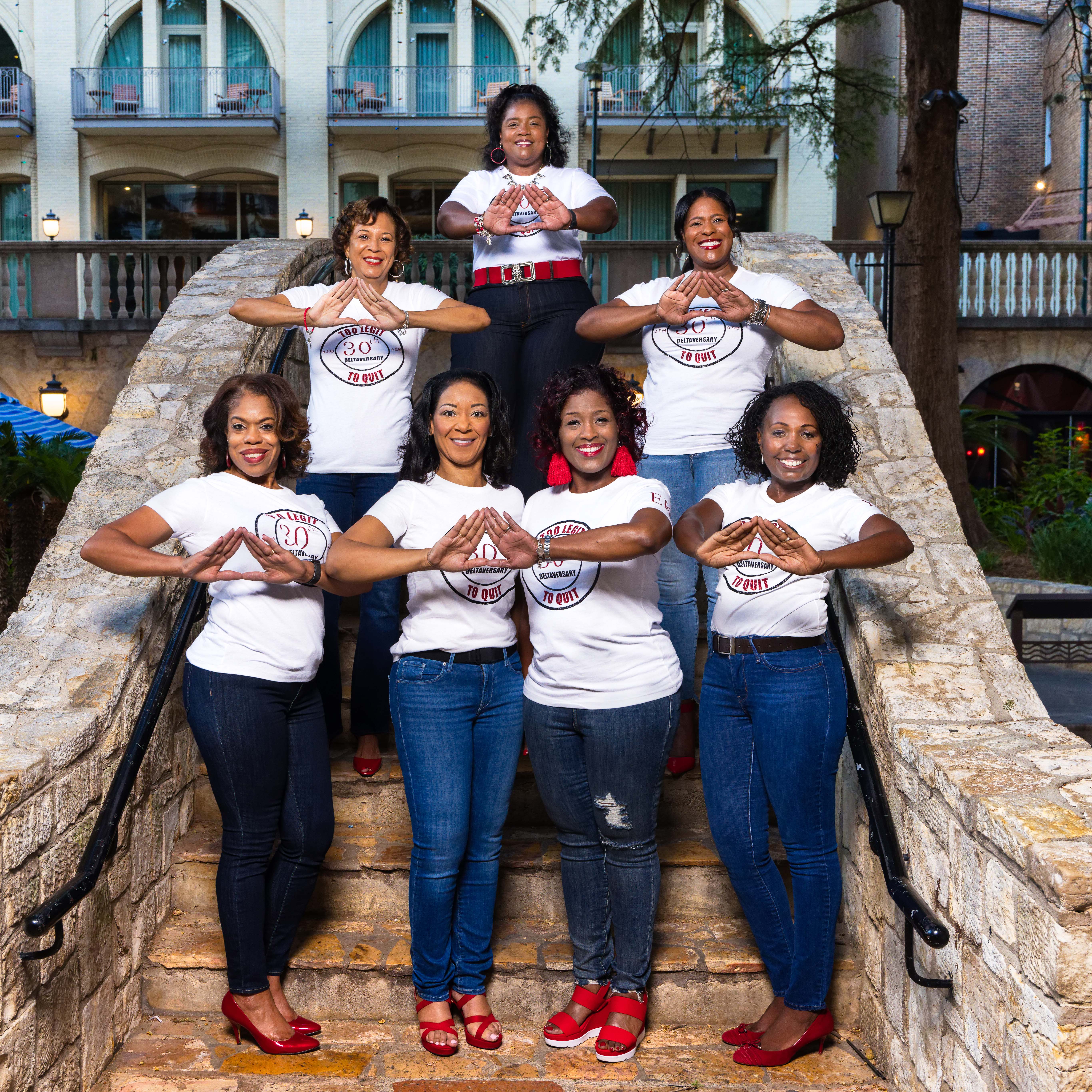 Seven women in white T-shirts and jeans forming diamond hand signs on stone steps outdoors.