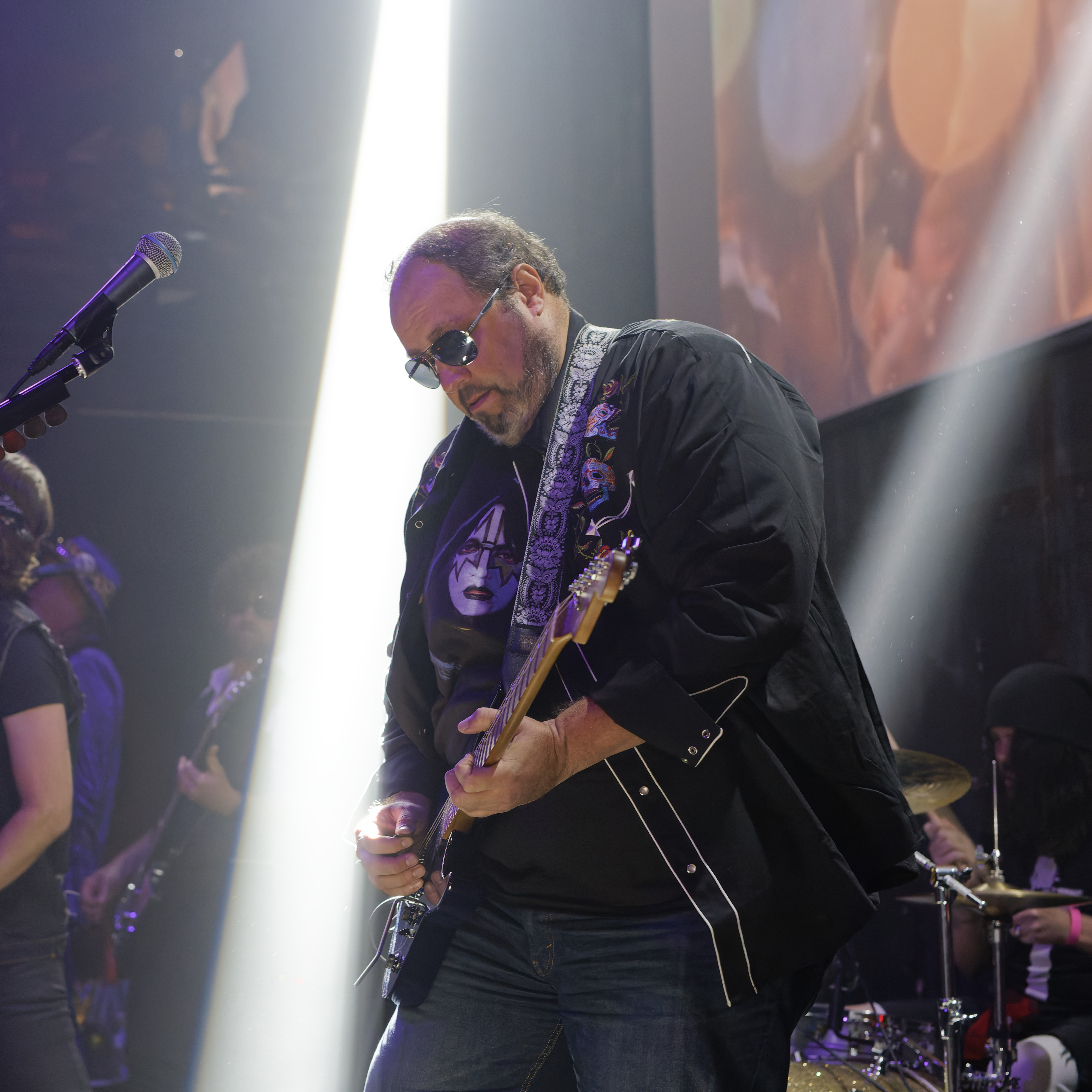 Man wearing sunglasses plays electric guitar on stage with drummer and band in background under spotlights.