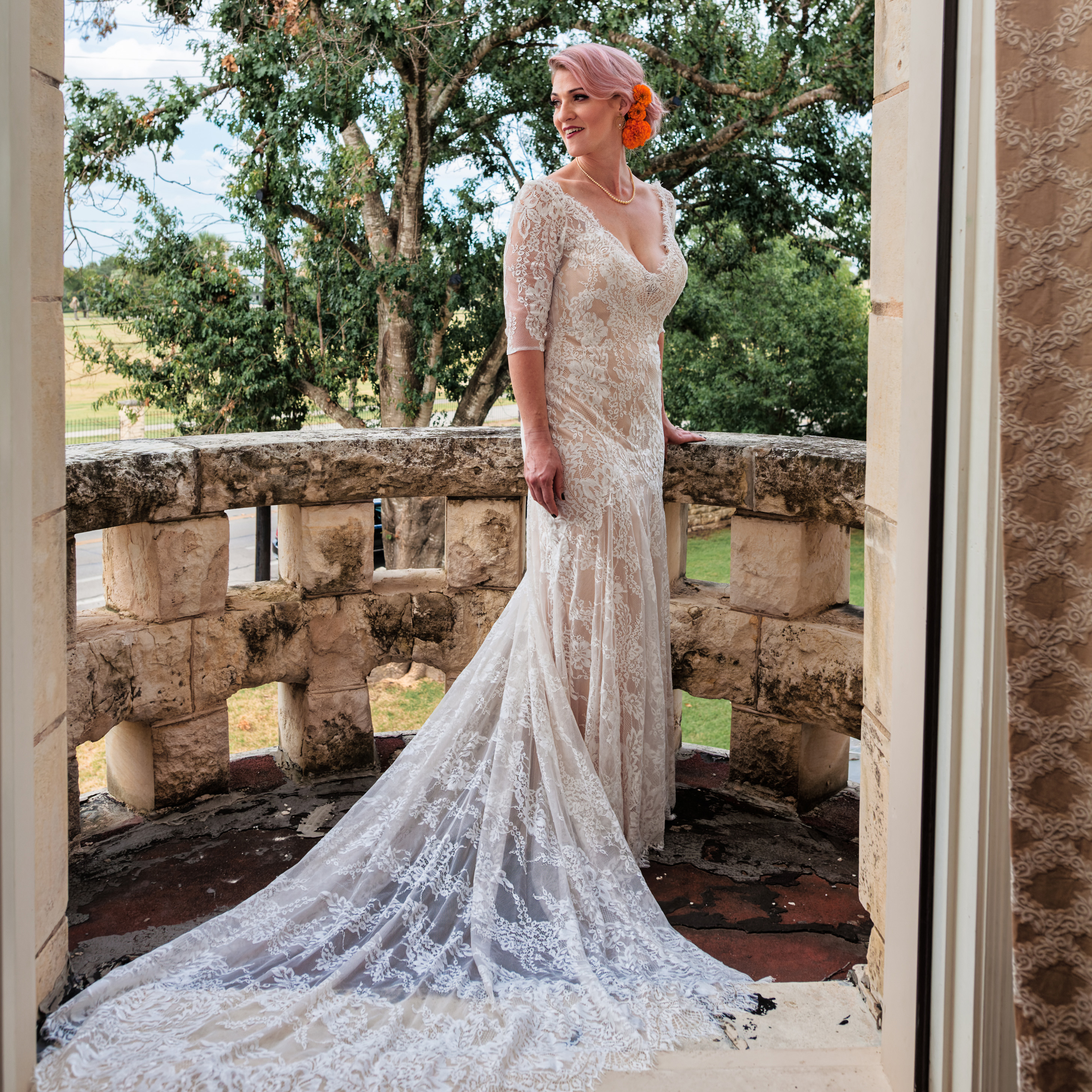 Woman in lace wedding dress with train stands on stone balcony with trees in background.