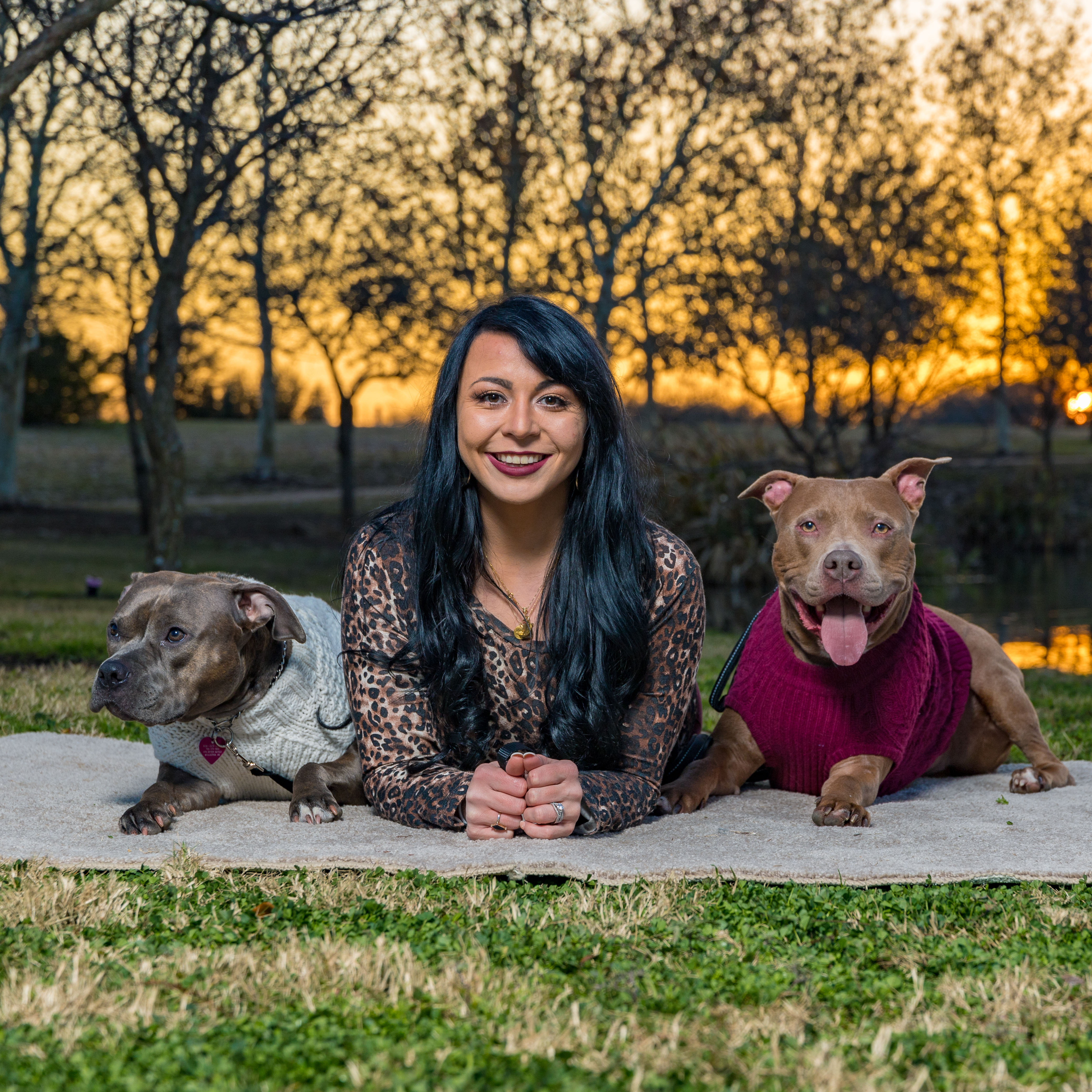 Smiling woman lying on a blanket outdoors with two dogs in sweaters during sunset.