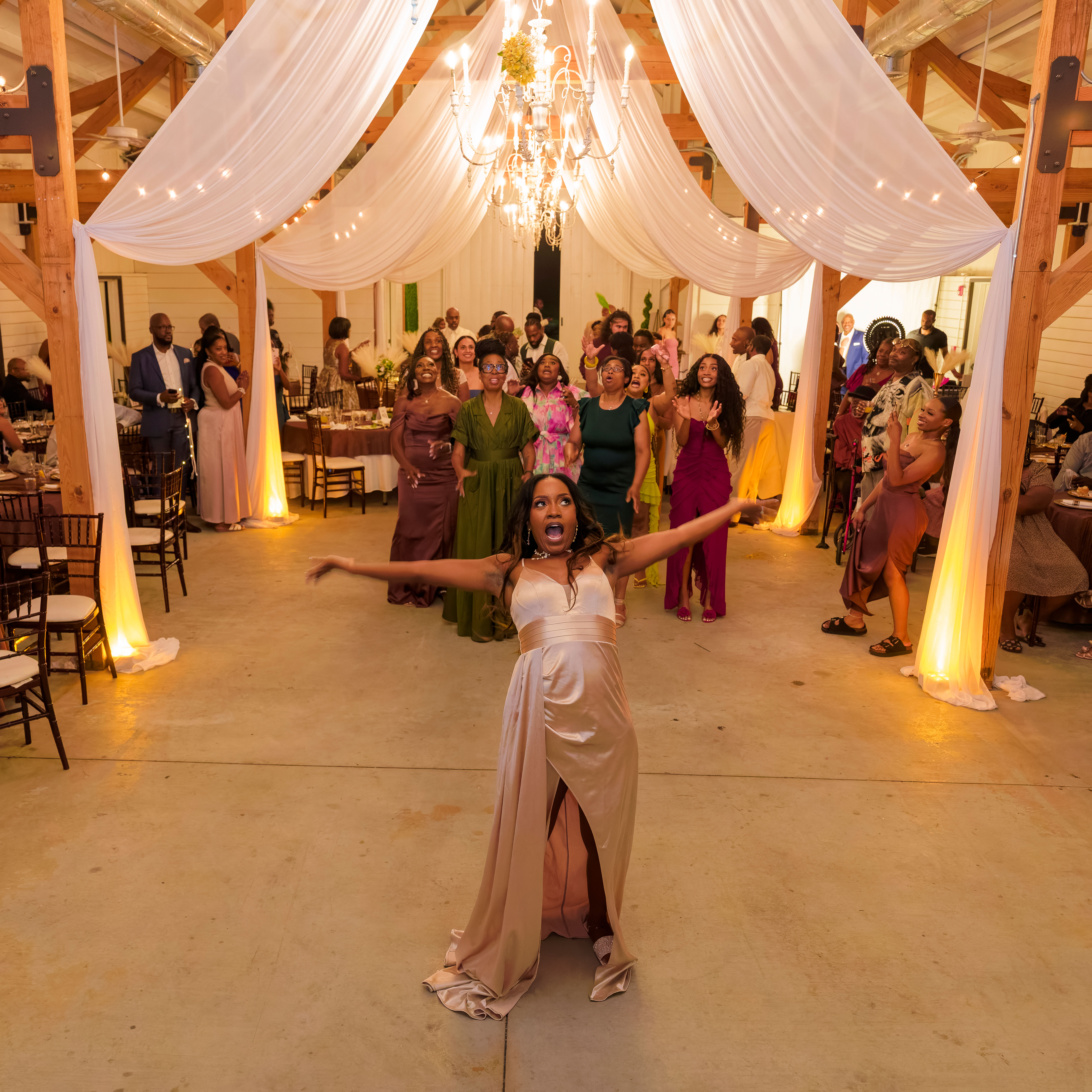 Woman in gold dress throws bouquet to eager group of women at elegant indoor wedding venue.