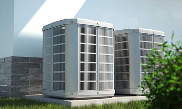 Two outdoor air conditioning units placed on a concrete slab next to a building wall with green grass and some shrubbery in the foreground.