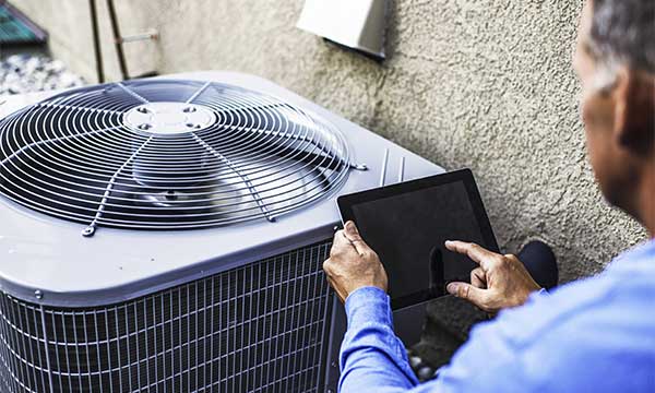 Technician using a tablet to inspect an outdoor air conditioning unit mounted against a textured wall.