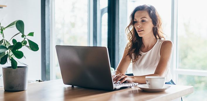Woman working on a laptop at a wooden table near a window with a cup of coffee and a potted plant.