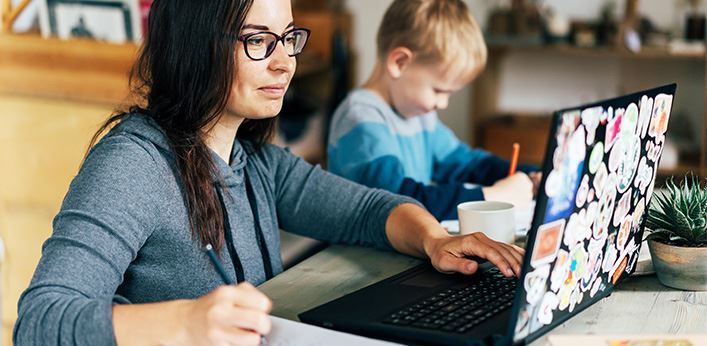 Woman working on a laptop covered with stickers while a young boy draws beside her at a table.