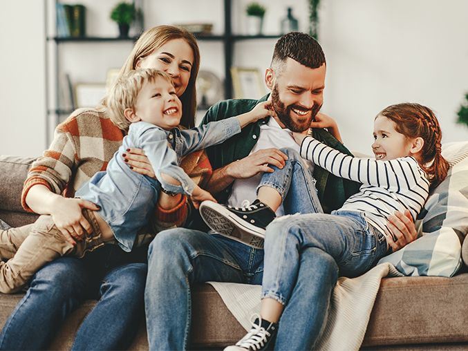 Smiling family of four playfully sitting on a couch with parents holding and tickling their two children.