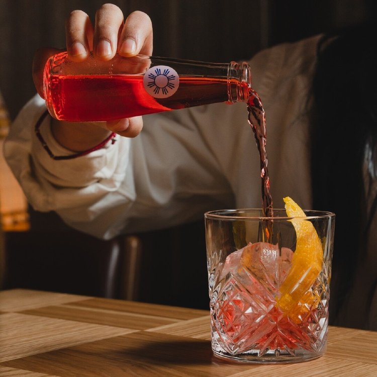 Hand pouring red liquid from a bottle into a glass with ice and an orange peel on a wooden table.